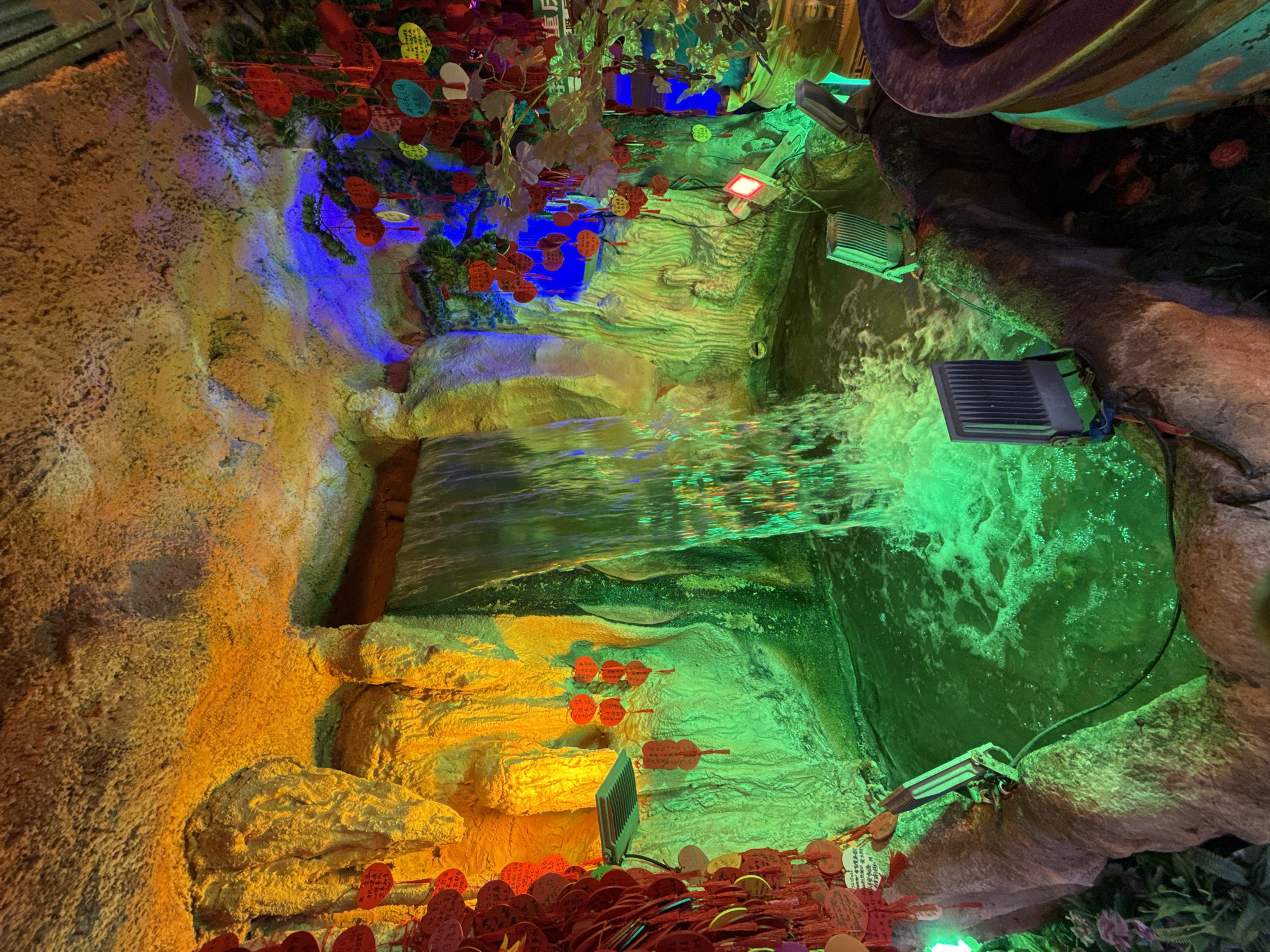 The image depicts a brightly illuminated indoor water feature, possibly a grotto or fountain. The walls are textured and colored in yellow, green, and blue. A waterfall cascades down, with several artificial lights directing green light onto the water and surrounding structures. Red and yellow paper ornaments are densely arranged near the bottom and hanging from above. Electrical lights and wires are visible, suggesting an artificial setup. The location is indicated as being in Yuzhong District, China.