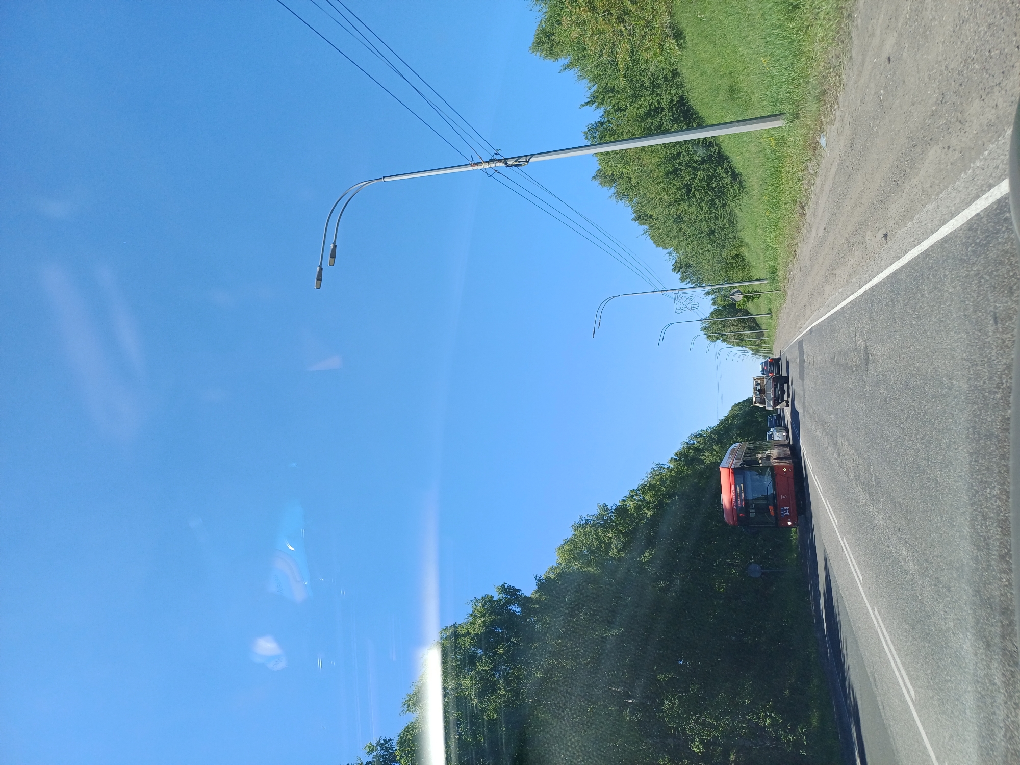 A bright, sunny day is depicted from the perspective of a vehicle driving on a two-lane road. Lush green trees line the left side of the road, and a grassy embankment with more trees flanks the right. A red bus is visible in the left lane, followed by other vehicles. Streetlights are spaced along the right side of the road, with power lines running overhead. The sky is a clear blue with some reflections visible on the vehicle's windshield. The road itself is paved and has white lane markings.