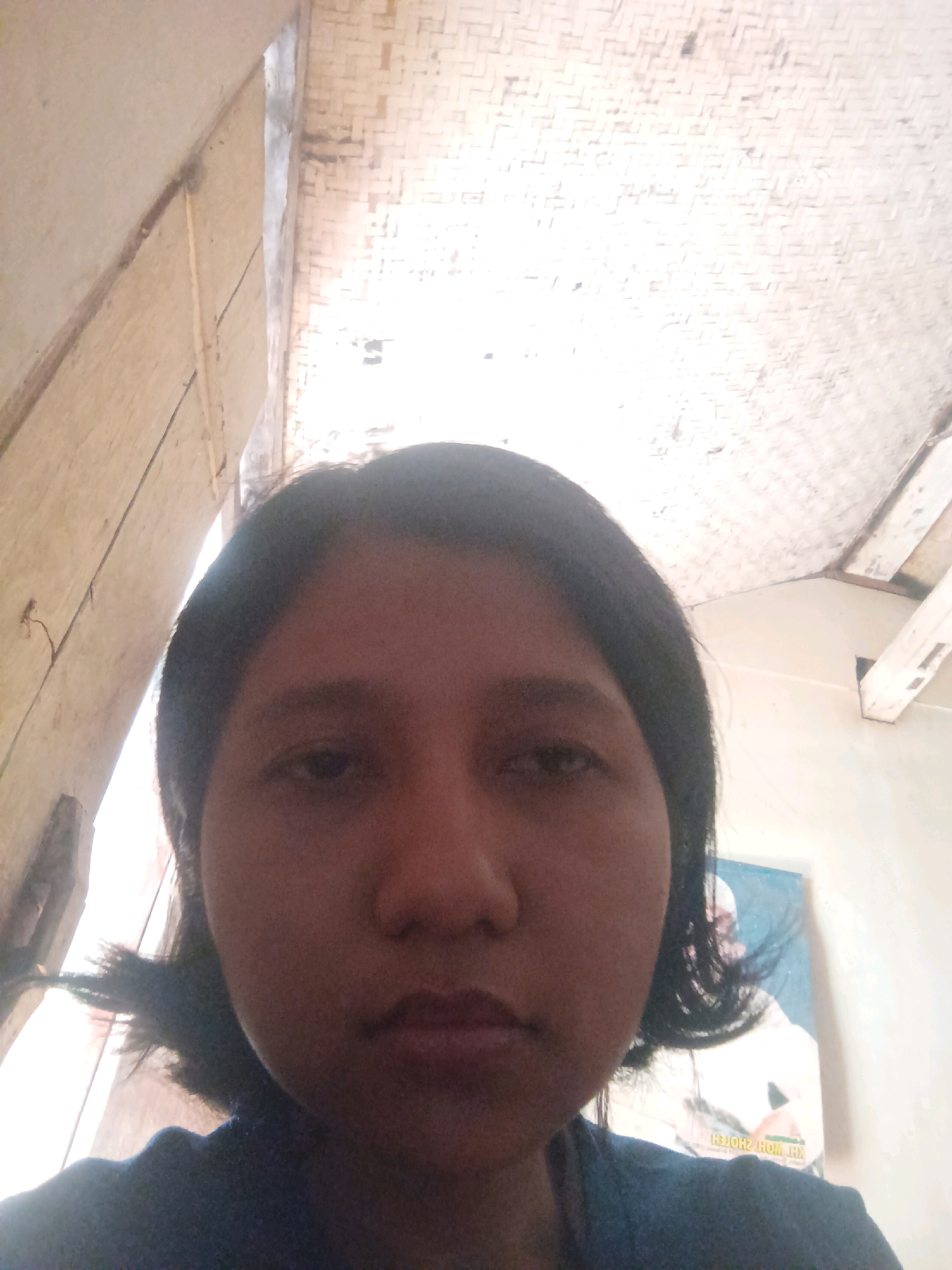 A medium shot shows a person with dark, shoulder-length hair in a dark blue shirt. The person is facing the camera with a neutral expression. In the background, a woven ceiling is visible, along with a picture of a person in white attire on the wall. The scene is indoors. The photo appears to be taken in Canga'an, Indonesia.