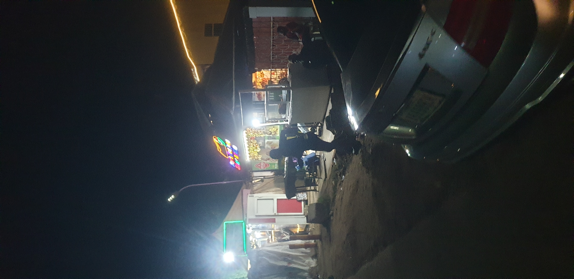 A nighttime street scene in Narayi, Nigeria, features several food stalls illuminated by various lights. In the foreground, the rear of a silver Mercedes-Benz sedan is visible, parked on a dirt road. The car has a license plate, though the characters are not clearly legible. To the left of the car, a narrow, unpaved road leads towards the stalls.
The food stalls are set up under makeshift shelters. One stall on the left has a green illuminated sign and appears to be selling something from a white structure with a red door. Further down, another stall features a neon sign that spells out "Royal King" with images of hot dogs and drinks. This stall is brightly lit with colorful string lights and is where a person in silhouette is seen working. To the right of this stall, a covered counter area displays food items. Further to the right, other individuals are seated, possibly customers or workers, in the dimmer lighting. The overall environment suggests a lively, albeit informal, nighttime commercial area.