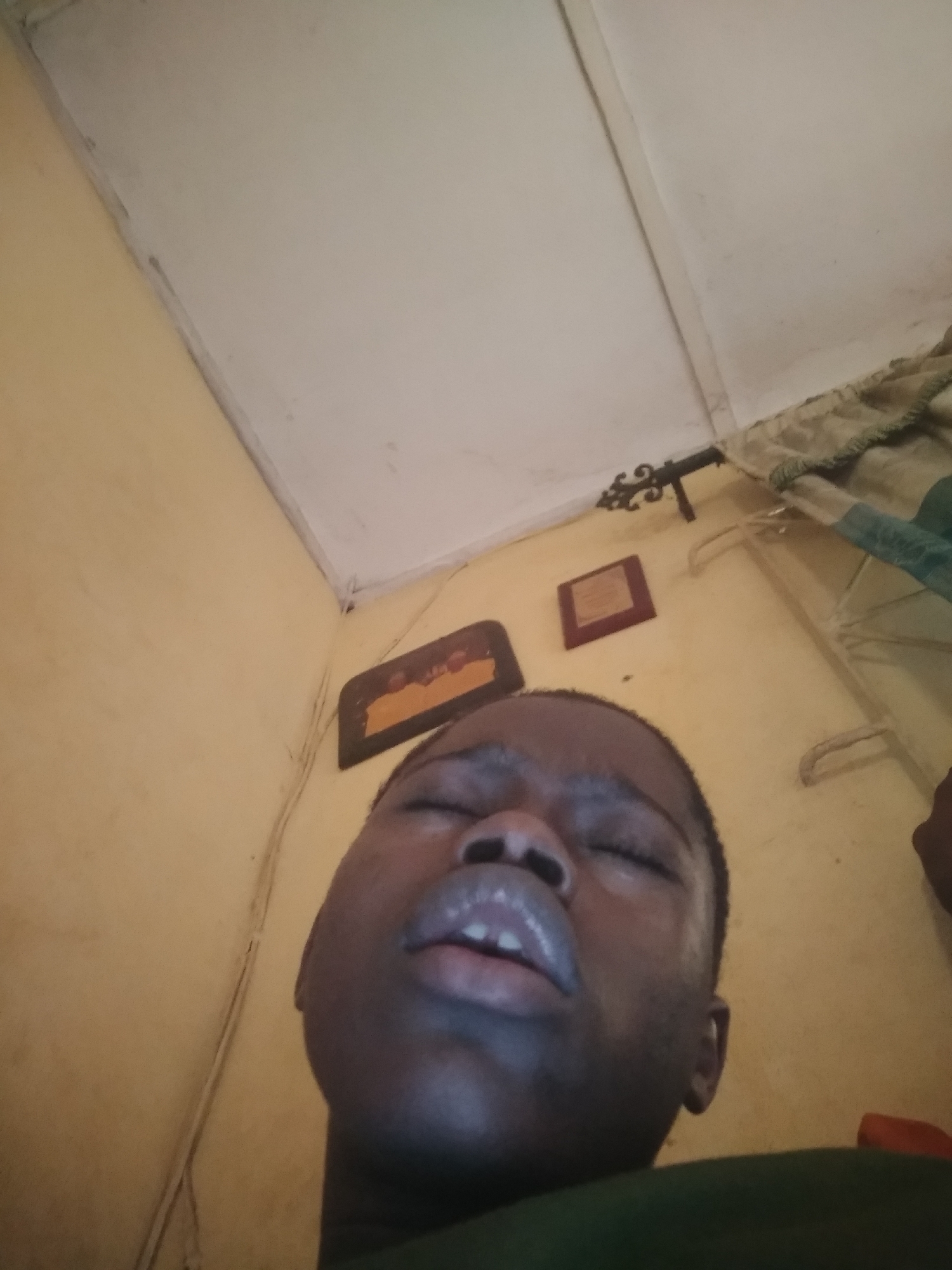 A low-angle, indoor shot shows a person lying down with their eyes closed and mouth slightly open. The person is dark-skinned. The ceiling above is white with trim. The wall behind the person is yellowish and features two framed pictures and decorative metalwork. A bed frame with bedding is visible in the top-right corner. Location is Unknown, Unknown.