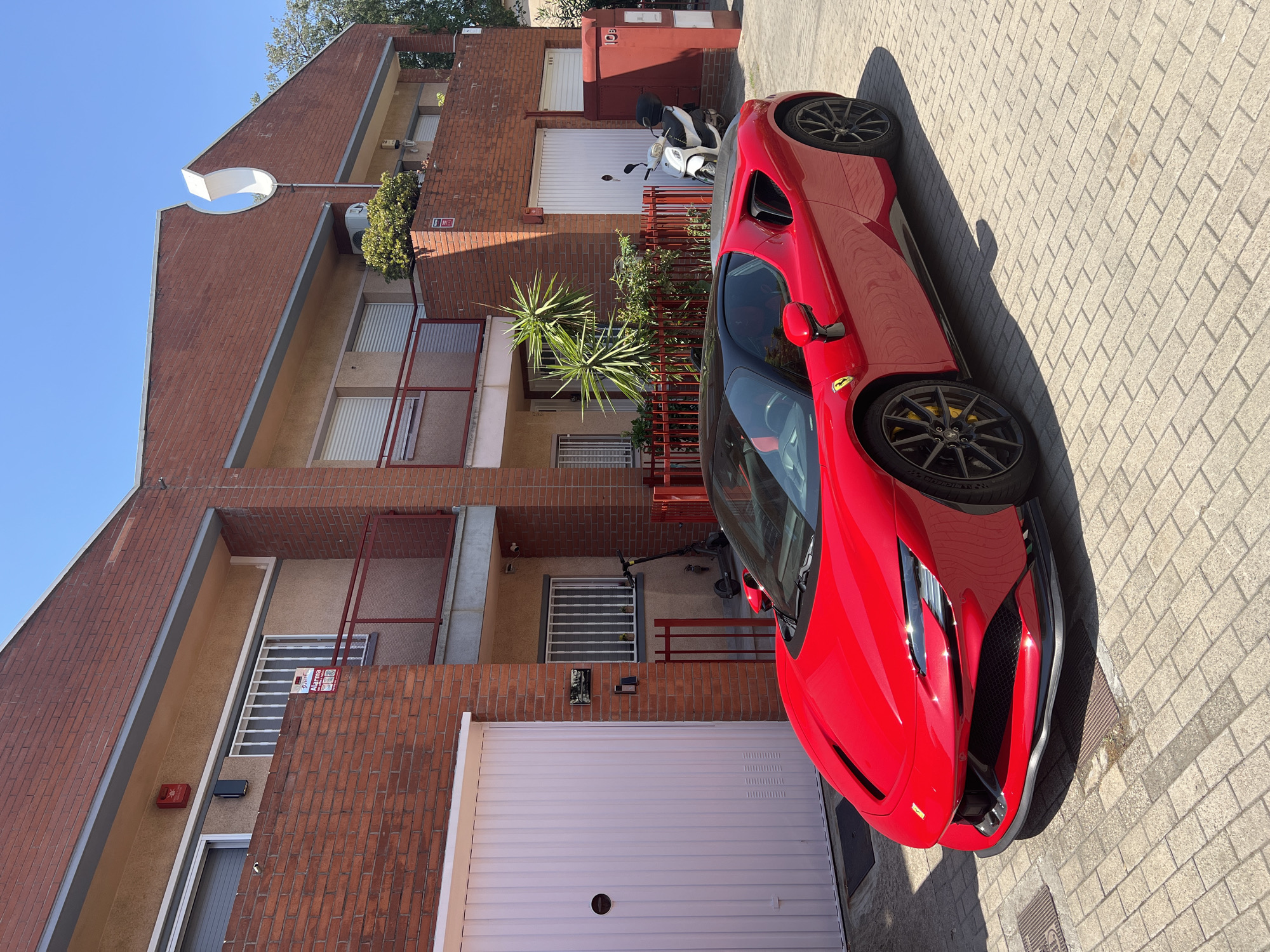 In the city of Mollet del Vallès, Spain, a vibrant red Ferrari sports car is parked on a brick-paved street in front of a two-story brick building. The building features light-colored stucco walls, multiple windows with white roller blinds, and red railings on balconies. A white garage door is visible on the left side of the building, and a smaller white garage door is on the right, next to a red mailbox or utility box. To the right of the main building, a white scooter is parked. The environment is bright and sunny, with a clear blue sky. An electric scooter is parked near a barred window on the lower level of the building, and a green spiky plant grows near the entrance. The Ferrari, a prominent subject, is angled towards the viewer, showcasing its sleek design and black roof. Its front hood features the iconic yellow Ferrari badge.
