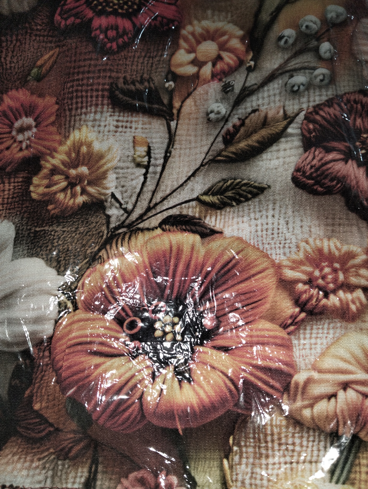 The image is a close-up, top-down view of a fabric with a floral pattern. The fabric appears to be textured, possibly linen or a similar weave, with a printed design of various flowers in shades of orange, yellow, brown, and red. Some of the flowers are large and in full bloom, while others are smaller or appear as buds. Delicate brown stems and dark green leaves are intertwined with the flowers, creating a naturalistic composition. A clear plastic sheeting is present, which has created a slightly distorted and glossy effect on the surface of the fabric. The lighting highlights the sheen of the plastic and creates reflections. The image does not contain any elements that would allow for the identification of a specific location within the city of Payagaboh, Indonesia, or any people, actions, or interactions.