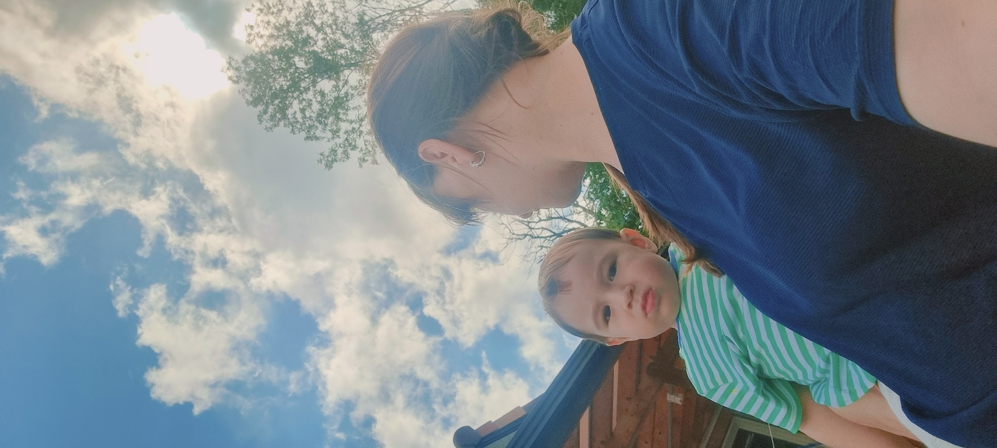 A low-angle shot captures a woman and a young child against a backdrop of a partly cloudy sky. The woman, positioned at the top of the frame, has her dark hair pulled back and is wearing a dark blue shirt. A small earring is visible in her left ear. The child, nestled in her arms, is wearing a green and white striped shirt. Behind them, the sky is a mix of blue and white clouds, with trees visible near the top. A section of a building with a dark roof can be seen at the bottom of the frame. The location is presumed to be Dunnae-myeon, South Korea.