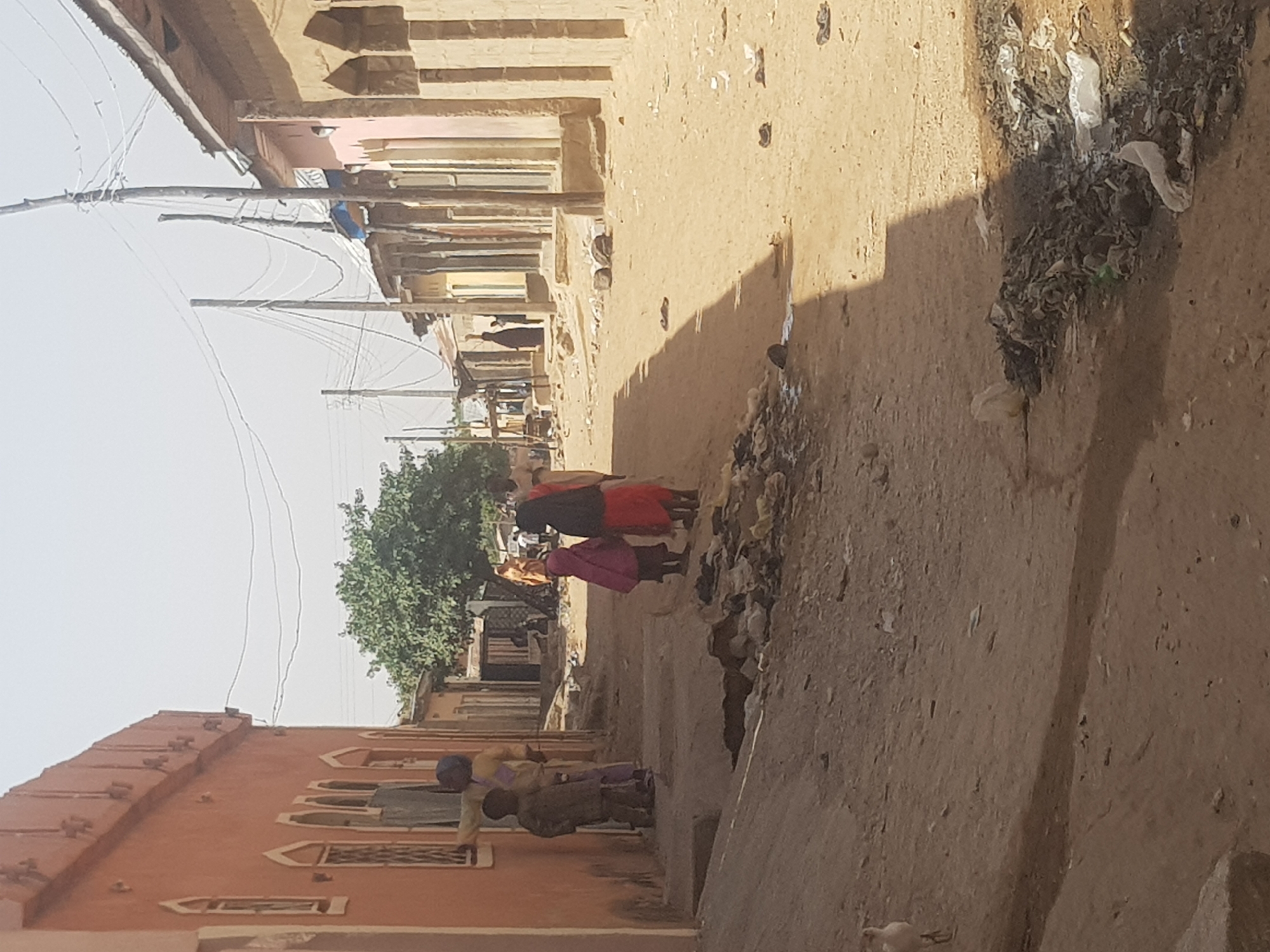 In Dabawa, Nigeria, the scene captures a narrow, dusty street lined with buildings. Two women are walking away from the viewer, while two children stand near a building. Debris and garbage accumulate along the roadside. Overhead, power lines are visible. The overall impression is of a simple, unpaved street in a residential area.