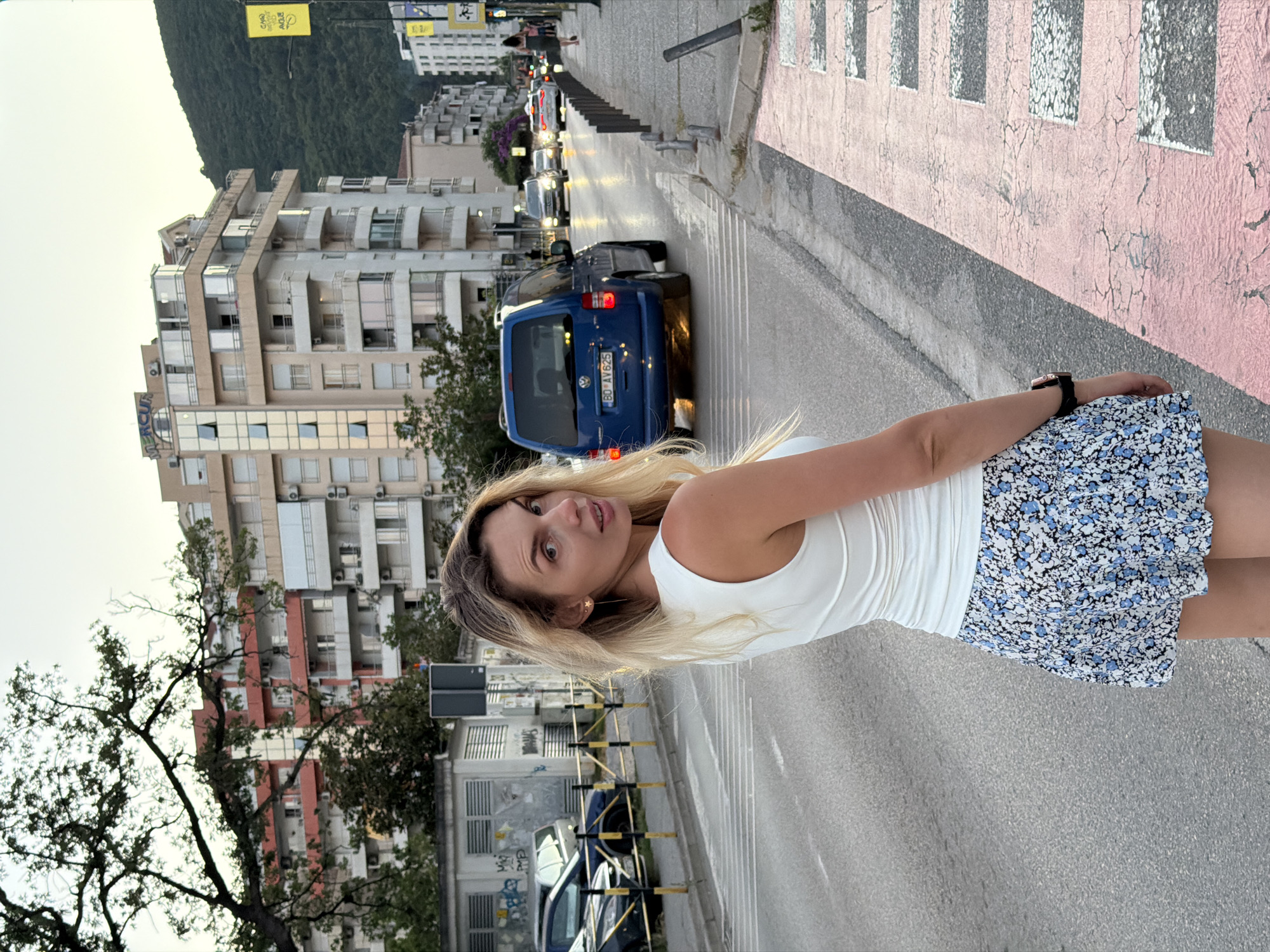 The image shows a woman standing on a pedestrian crossing in Budva, Montenegro. She is looking over her shoulder at the camera, and she is wearing a white tank top and a floral skirt. In the background, there are cars driving on the street, and buildings on either side of the street. The sun is setting, and the sky is orange and pink.