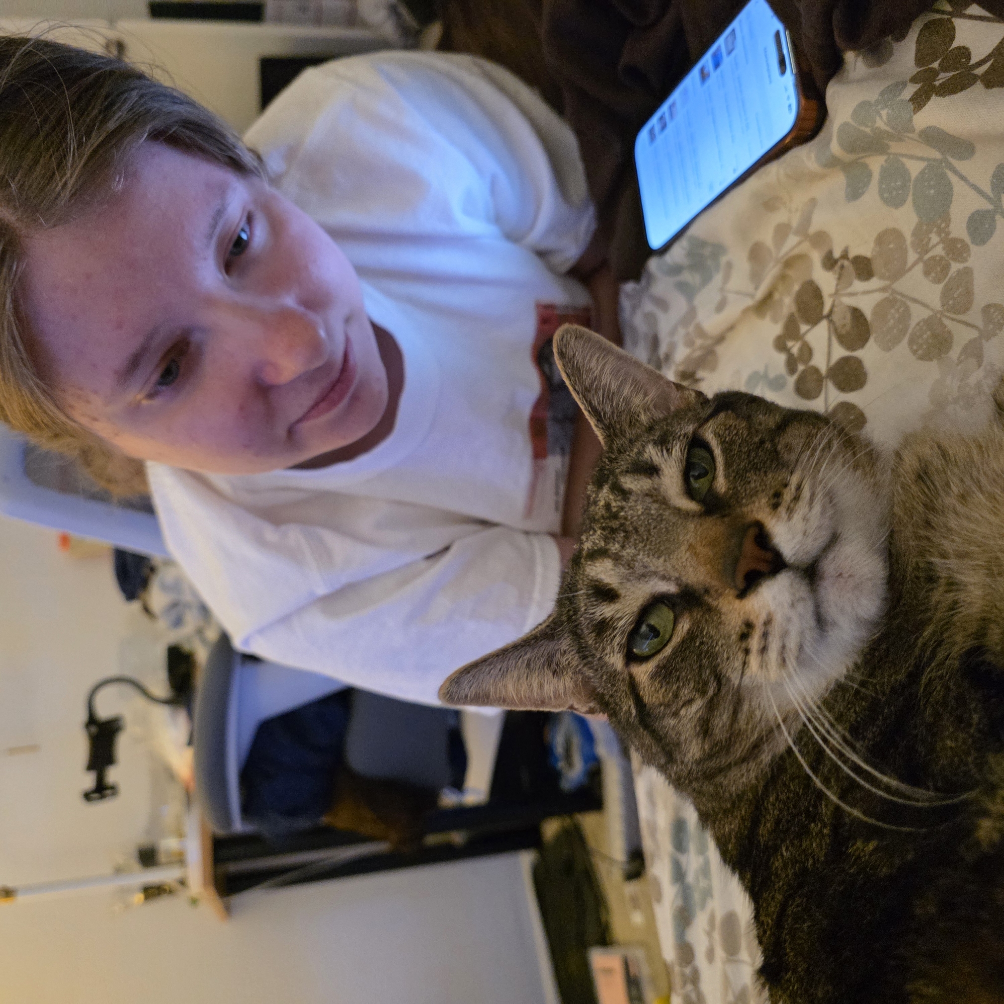 In Portland, United States of America, a close-up, eye-level shot shows a tabby cat resting on a patterned bedspread. The cat's face, predominantly in the foreground, is a mix of brown, black, and white fur with striking green eyes. To the right and slightly behind the cat, a person with light-colored hair and fair skin is visible from the shoulders up, wearing a white t-shirt. The person is looking down, seemingly at a smartphone with a bright blue screen that rests on the bedspread to the right of the cat. The background is blurred but suggests an indoor setting with a desk, chair, and a lamp, typical of a bedroom or living space.