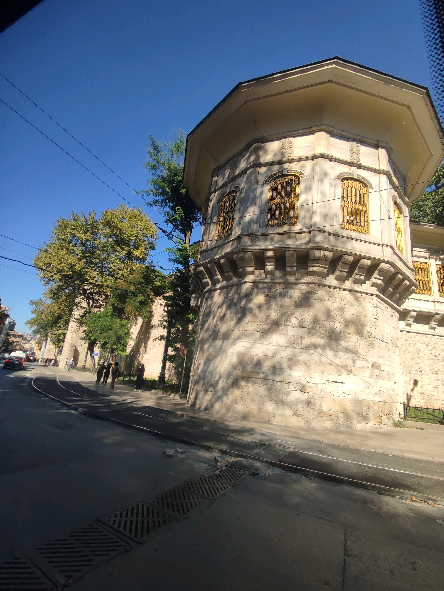 An octagonal stone building with yellow-grated windows stands on a street corner in Istanbul, Turkey, on a clear, sunny day. The building is adorned with ornate stonework and a wide, overhanging roof. A large tree with green foliage grows beside it, casting shadows on the building's facade. A street with tram tracks runs in front of the building, and several pedestrians are walking on the sidewalk. The street appears to be under construction or repair, with visible manhole covers and some debris. The sky is a vivid blue, with power lines strung across it. The overall scene suggests an urban environment with historical architecture.