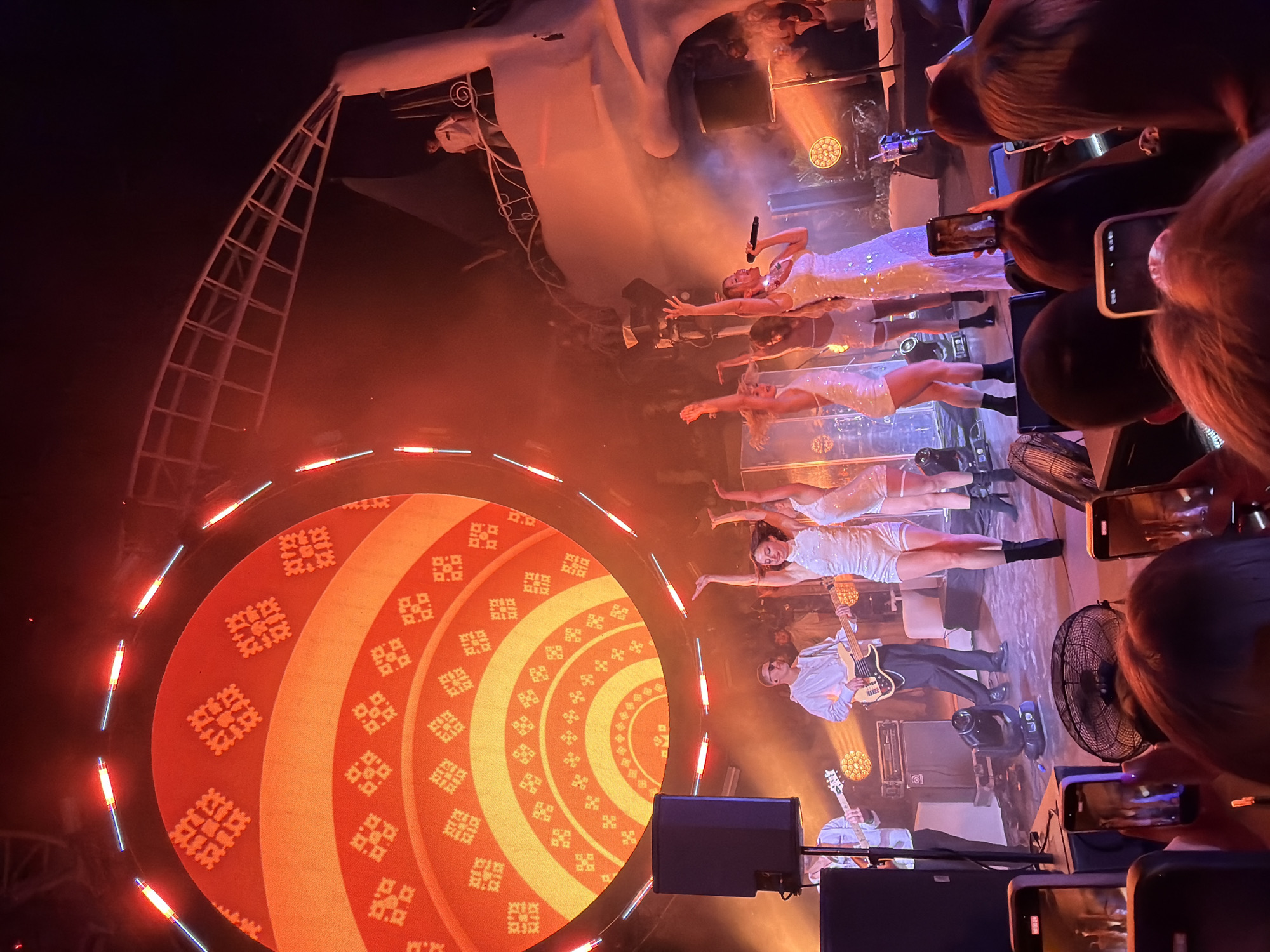 The image depicts a vibrant concert scene on a stage with a large, circular LED screen displaying an orange, geometric pattern. A group of female performers in sparkling white outfits are on stage, with one singer holding a microphone. They appear to be dancing and interacting with the audience. Two male musicians, one playing a bass guitar and the other a regular guitar, are also on stage.
In the foreground, the backs of several audience members are visible, many of whom are holding up smartphones to record the performance. The lighting is dramatic, with warm orange and red hues dominating the scene, creating a dynamic and energetic atmosphere. Smoke or haze is present on stage, enhancing the lighting effects. The overall impression is of an exciting live music event.
The concert appears to be taking place in the city of Odesa, Ukraine.