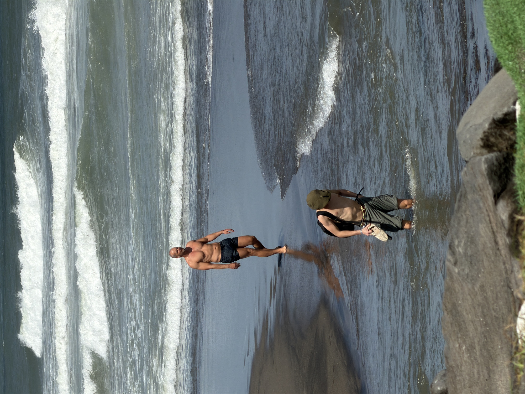 The image captures a beachfront scene in Mengwi, Indonesia, with two men present. In the mid-ground, a bald man with a muscular physique walks on the wet sand near the shoreline, wearing dark swim trunks. Further in the foreground, another man, partially obscured, stands in shallow water. He is wearing olive-colored cargo shorts and a backpack, and holds a sandal in his right hand. The ocean behind them displays multiple rows of white-capped waves rolling towards the shore under a bright sky. The beach itself appears to be composed of dark sand. Large rocks are visible in the lower foreground, with some green vegetation.