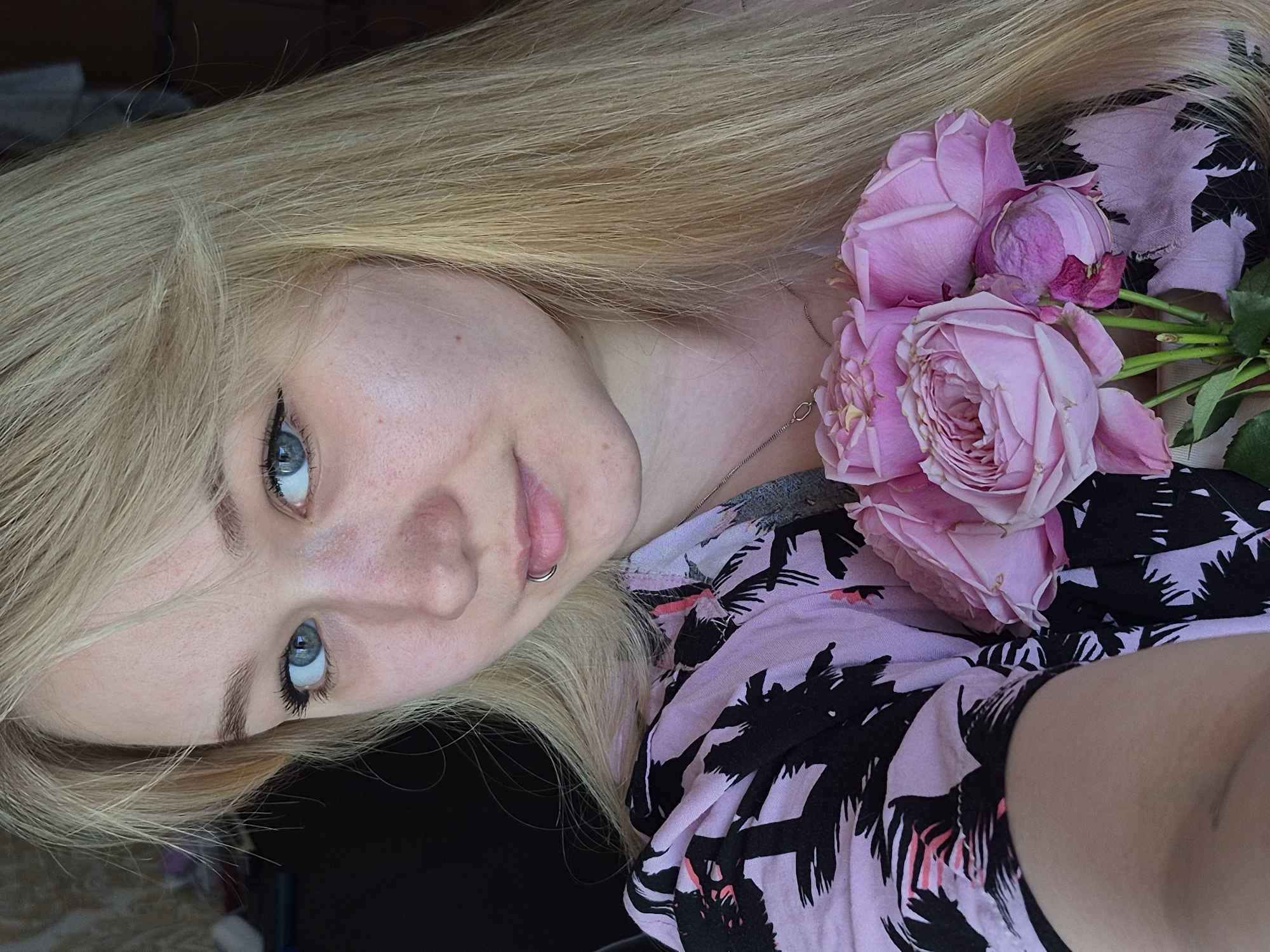 A fair-skinned, blue-eyed young woman with long blonde hair, wearing a light pink shirt with a black palm tree print, is shown in a close-up, slightly angled view. She has a lip ring on the left side of her mouth. A bouquet of pink roses rests on her chest. The location and city of Chaikovsky, Russia, are not discernible from the image.