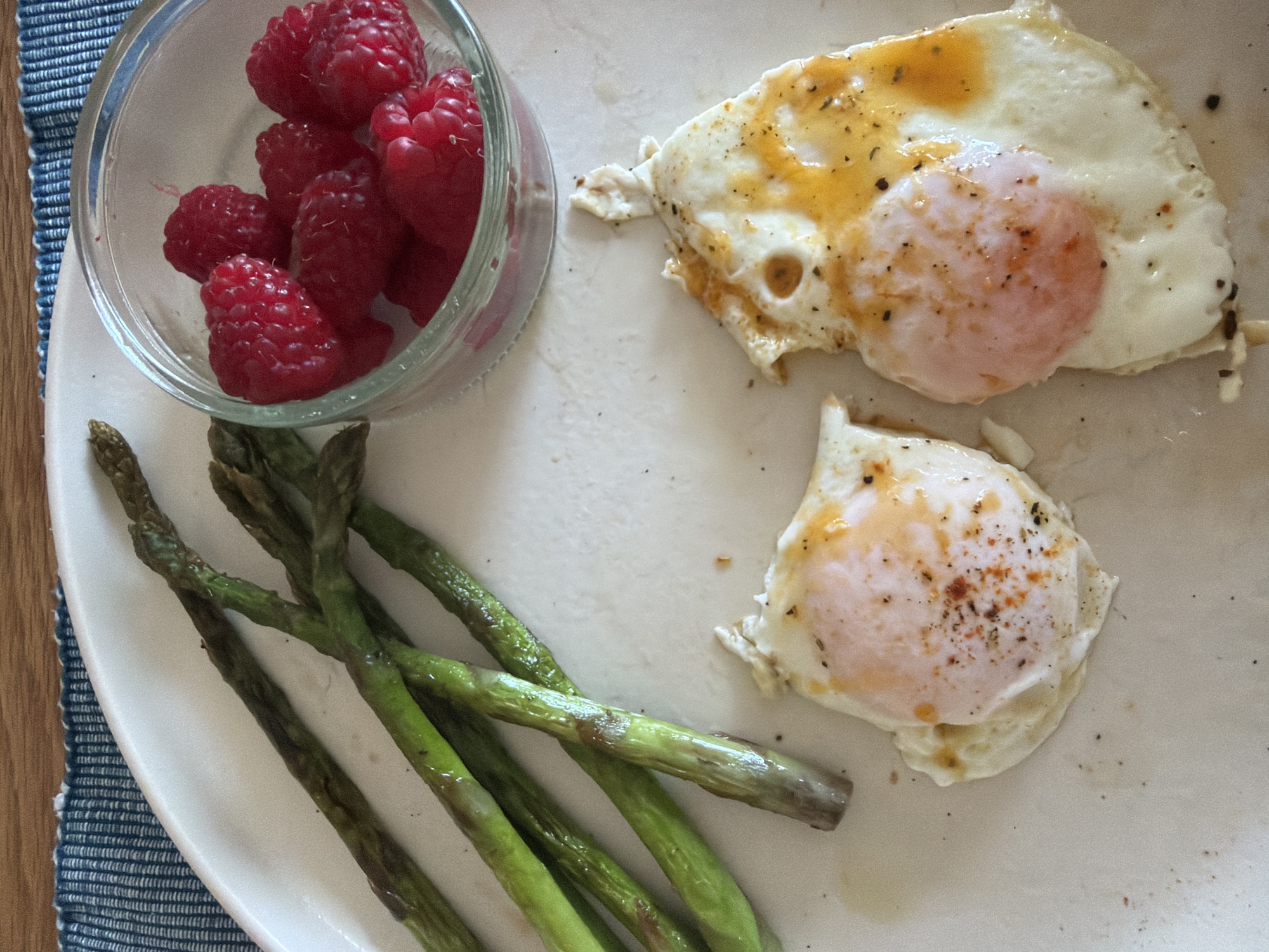 A top-down shot of a white oval plate displaying a meal consisting of raspberries in a small glass bowl, a cluster of cooked asparagus spears, and two fried eggs. The raspberries are bright red, clustered together in the bowl. The asparagus spears range in color from dark green to a lighter, slightly yellowish green, suggesting they have been cooked. The fried eggs have cooked whites and runny yolks, seasoned with pepper. The plate sits on a blue and white woven placemat. The presence of the placemat suggests a domestic or restaurant setting. It is not possible to ascertain that this photo was taken in Sofia, Bulgaria, without further contextual information. There is no indication of people in this photo.