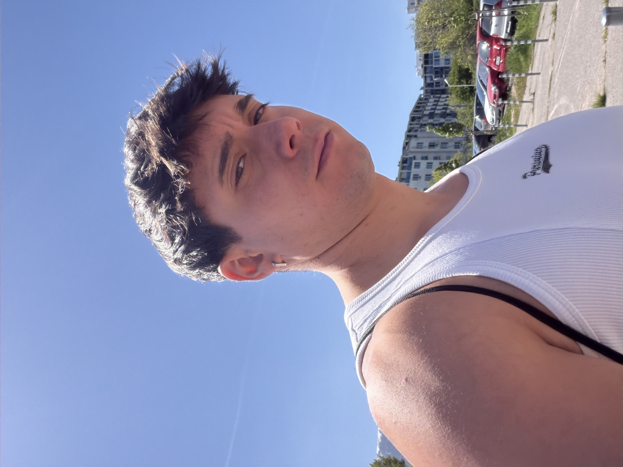 A low-angle, medium shot depicts a fair-skinned young man with short, dark hair and a slight tan, wearing a white ribbed tank top with a small logo on the right breast. The man is outside with a clear blue sky behind him. In the background are several parked cars, a building and some trees. The building is located in Sofia, Bulgaria. The man is looking down into the camera. He has an earring on his left ear.