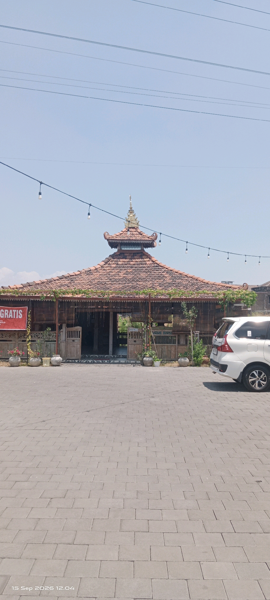 The image depicts a traditional Javanese building with a tiered, red-tiled roof, situated outdoors on a paved area. The building's facade is made of wood and features intricate carvings and decorative elements. A prominent red banner with white text reading "GRATIS" is visible on the left side of the building. Several potted plants with flowers are placed in front of the structure. A white MPV is parked on the right side of the paved area. Overhead, power lines crisscross the clear blue sky, with several decorative light bulbs hanging from one of them. The timestamp "15 Sep 2026 12.04" is present in the bottom left corner of the image. The scene appears to be a commercial establishment or a cultural site. The location is identified as Pabelan, Indonesia.