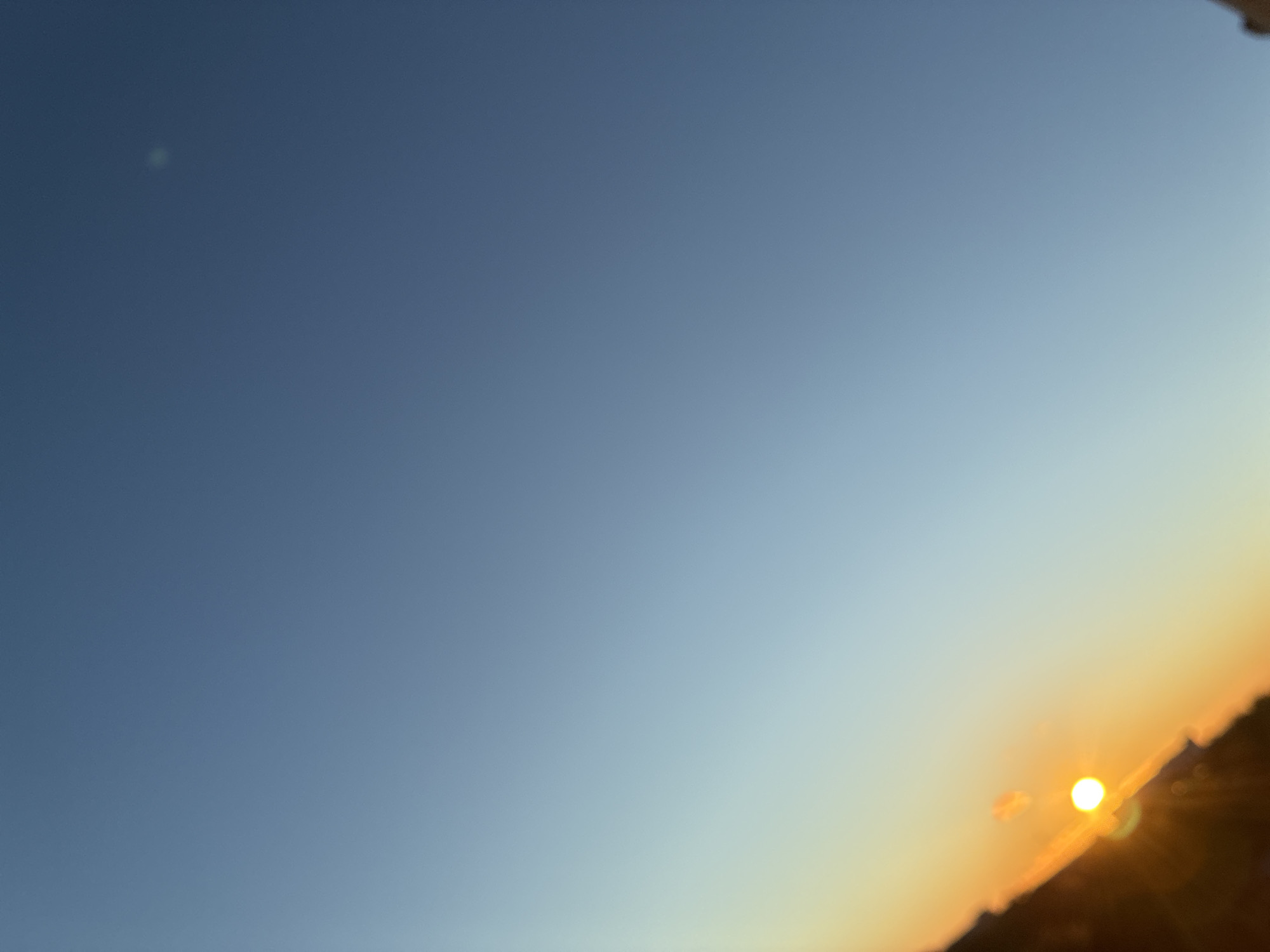 This is a landscape photograph of a sky at dusk. The sky transitions from a deep blue at the top of the frame to a pale yellow and orange near the horizon. A bright, circular sun is visible low in the frame, casting a warm glow and creating lens flares. The horizon is obscured by dark, silhouetted shapes that appear to be the tops of buildings or hills. There is a faint, pale green or white smudge in the upper left quadrant of the sky, possibly a cloud or atmospheric phenomenon. The overall impression is one of a serene and peaceful sunset. The time of day is clearly dusk, given the colors of the sky and the low position of the sun. The weather appears to be clear, allowing for a vibrant sunset. The location is unknown but appears to be an urban or semi-urban environment based on the silhouetted structures. No people or text are visible in the image.