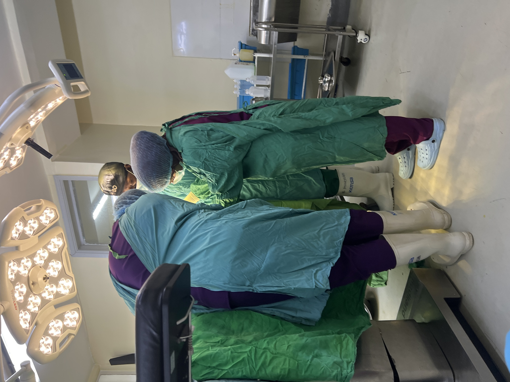 In what appears to be an operating room, two individuals clad in surgical attire are positioned over a patient who is not visible. The surgical staff are wearing protective green surgical gowns and boots. One is wearing white Crocs. A surgical light fixture illuminates the scene from above. A stainless steel surgical cart is visible in the background. The image was likely taken at a hospital or clinic in Nairobi, Kenya.