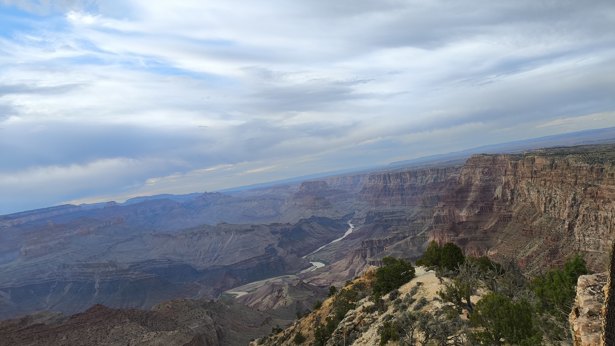 This is a panoramic, eye-level, landscape photograph of the Grand Canyon in Grand Canyon Village, Arizona, United States. The view looks eastward into the canyon, with the Colorado River winding through the bottom. The sky is filled with dramatic clouds, ranging from light blue to dark gray, indicating a dynamic weather system. The canyon walls are immense and multi-layered, displaying a spectrum of earthy tones from reddish-brown to muted grays and purples, sculpted by erosion into intricate formations.
In the foreground, the edge of the canyon rim is visible, covered in sparse desert vegetation, including dry grasses, shrubs, and a few small, gnarled trees. The terrain is rocky and uneven. The middle ground features the vast expanse of the canyon, with its receding layers of rock formations and the visible ribbon of the river. In the distance, the canyon stretches out, with hazy, mountainous horizons fading into the atmospheric perspective. There are no people or animals visible in the image. The overall impression is one of immense scale, natural beauty, and geological grandeur.