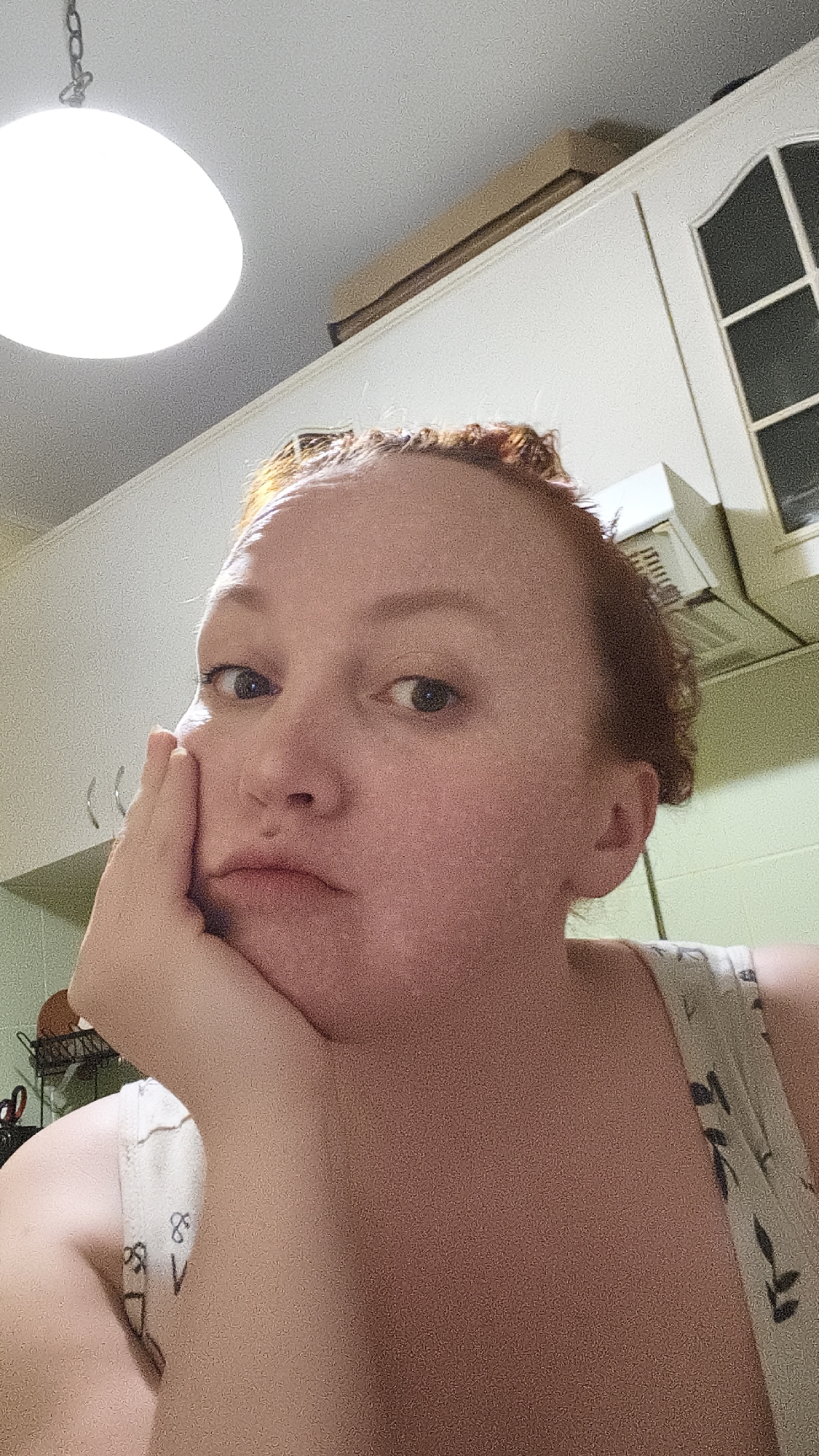 A reddish-haired person with their face resting on their hand is in what appears to be a kitchen. Behind them are white cabinets, a window with multiple panes, and an overhead light fixture. A box is resting on top of the cabinets.