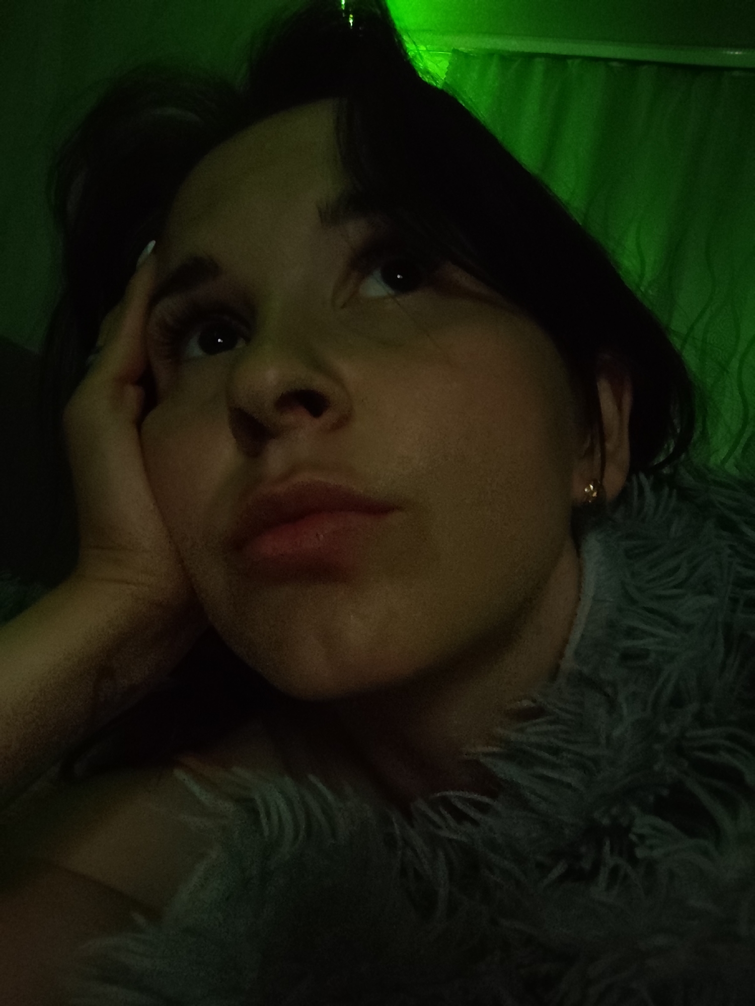 A person is resting their head on their hand, looking upward and slightly to the right. They have dark hair and fair skin, with an earring visible on the right ear. The person appears to be wearing a furry, grey-blue garment. The background is a green wall or curtain. The image has a green tint. It is not possible to determine if the location is Фоки, Russia from the image.
