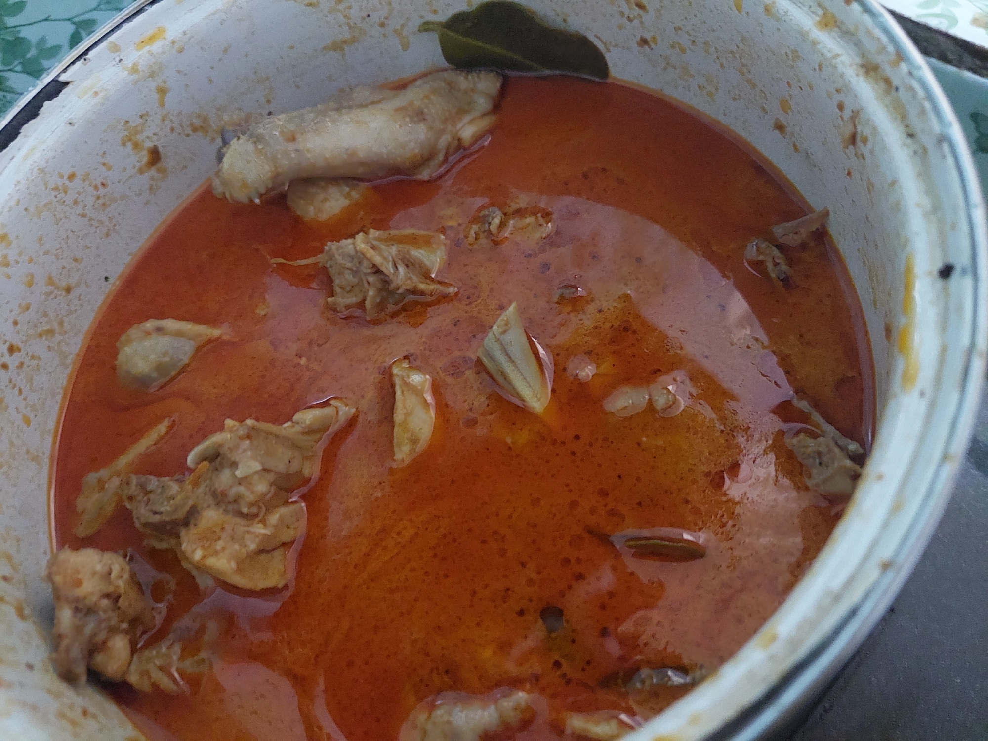 A close-up, overhead view shows a white pot filled with a rich, orange-red curry containing pieces of chicken. The curry is oily, with some visible spices and herbs. The pot itself is slightly stained and worn, suggesting it has been used frequently. A green leaf, likely a bay leaf, is visible in the curry on the right side of the pot. The background is blurred, but appears to be a table surface with a faint green and white patterned tablecloth. There is no indication of the city of Jelutung, Indonesia in the image.