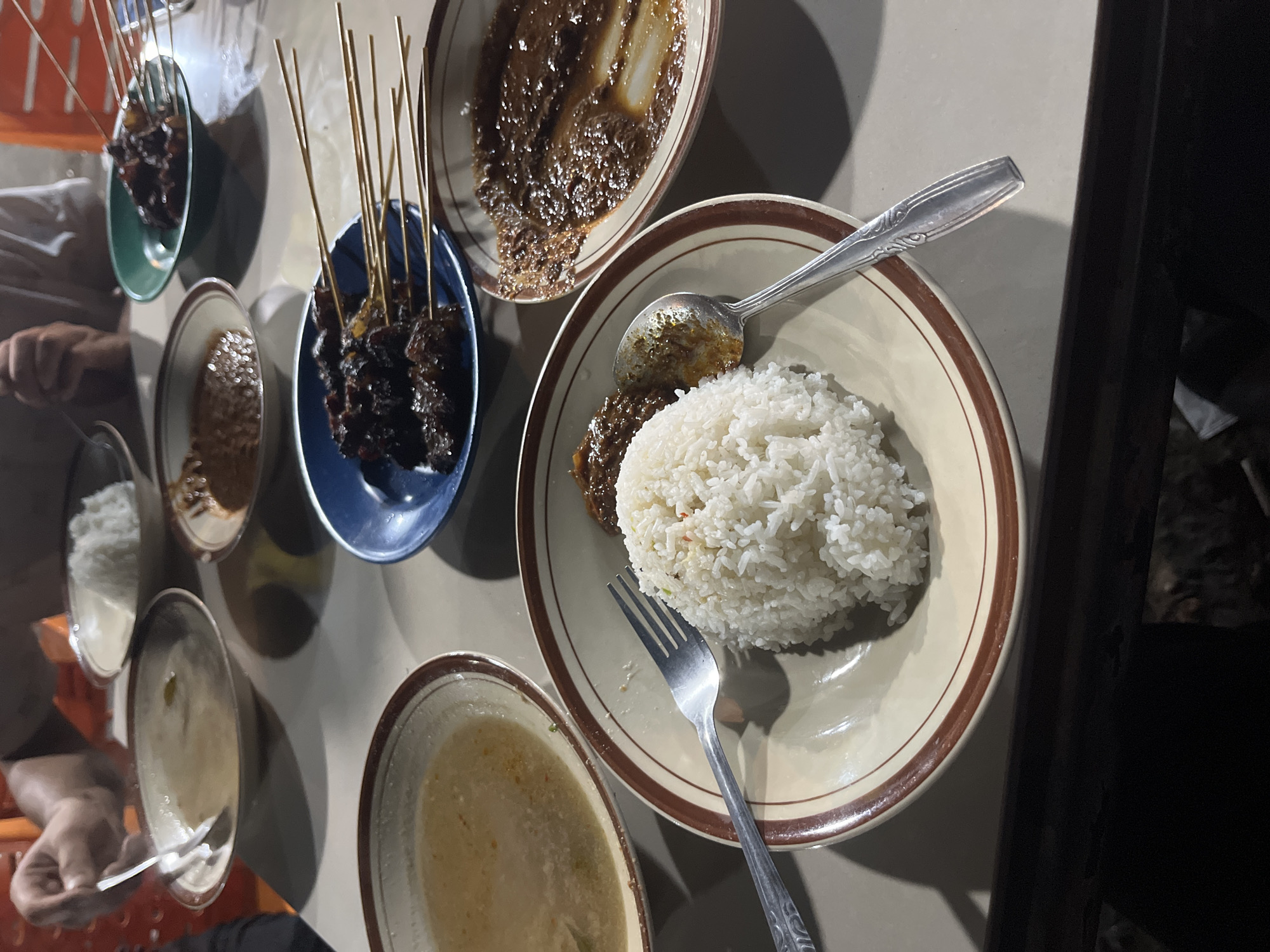 In Cotpuuk, Indonesia, this image captures a close-up view of a table laden with various dishes, likely part of a meal. The setting appears to be a casual dining establishment or a home. In the foreground, a plate holds a mound of white rice, accompanied by a fork and a spoon. To the left, a bowl contains a light-colored soup with some green flecks. Adjacent to these, another plate features a serving of dark, saucy meat, possibly beef or lamb. Further in the background, a blue bowl contains several skewers of grilled meat, likely satay, served with a side of a dark, thick sauce. Another green bowl holds a portion of what appears to be a savory dish. The table itself is a light-colored surface, with subtle lighting suggesting an indoor evening setting. The presence of multiple dishes and utensils indicates a shared meal among individuals, though only a partial view of a person's hand and arm, holding a fork, is visible in the upper left corner, engaging with the soup. The overall impression is one of a communal dining experience in a local Indonesian setting.