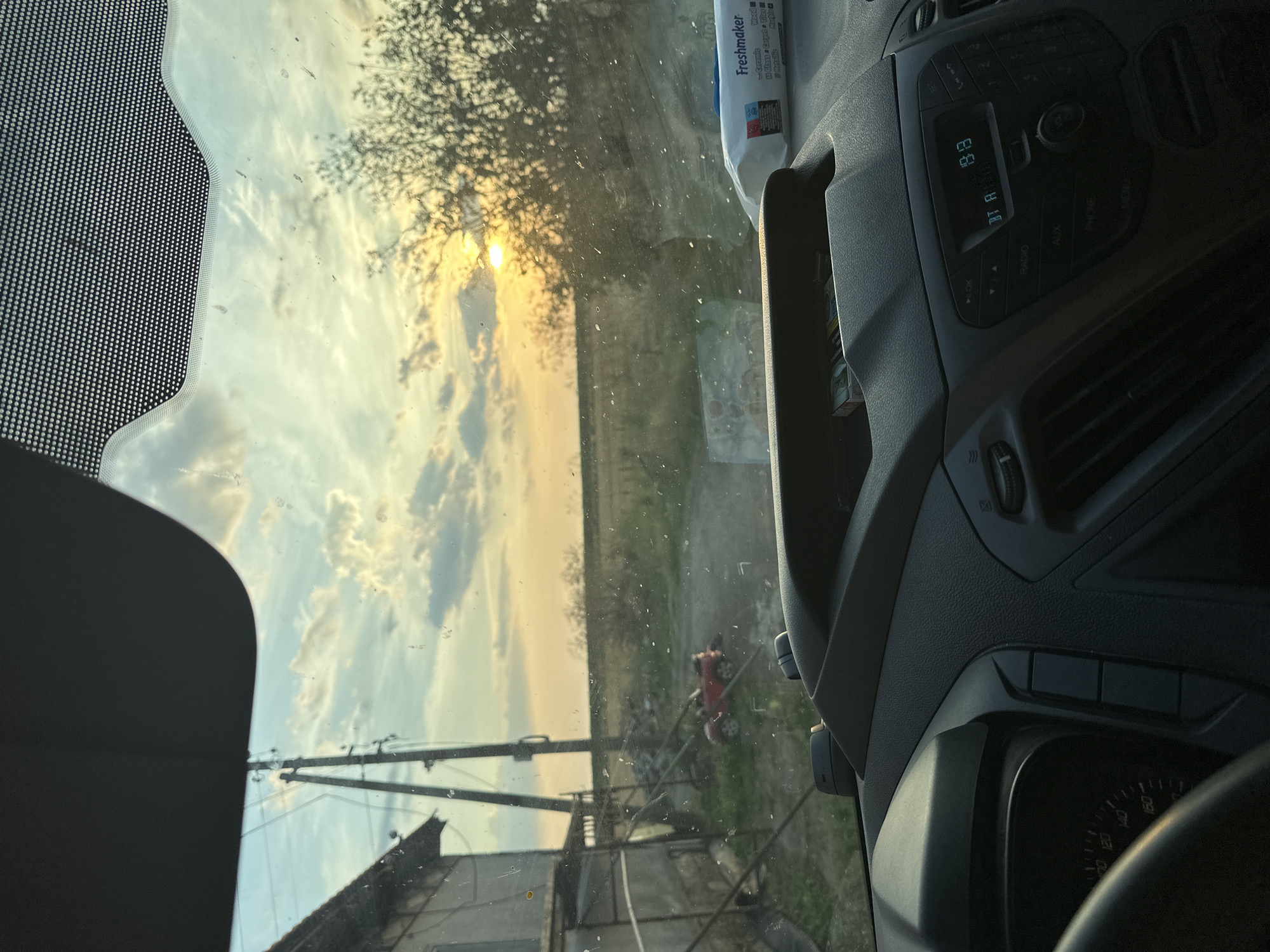 The image, taken from inside a vehicle, captures a sunset scene in Mlekarevo, Bulgaria. The view is framed by the car's interior, including the dashboard and windshield. The windshield is speckled with dirt or water droplets. Outside, a rural landscape unfolds with a house and a utility pole on the left, a dirt road leading into the distance, and trees on the right. A small red toy car is visible on the dirt road. The sun is setting, casting a warm glow through the trees and clouds in the sky. On the dashboard, a packet of "Freshmaker" wet wipes is placed next to a cigarette pack. The car's radio display shows the time as 19:19.