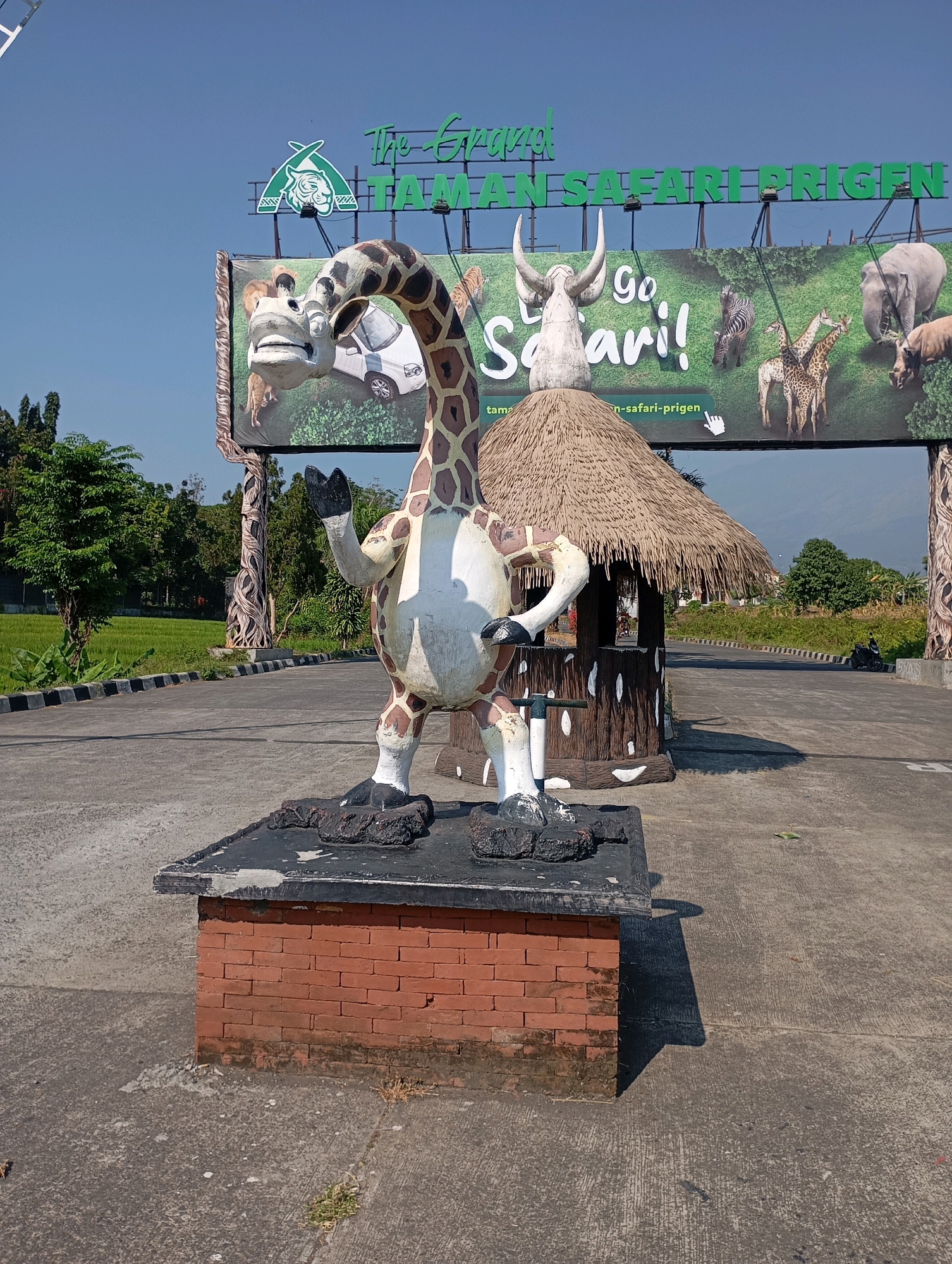 This image captures the entrance to Taman Safari Prigen, a zoological park located in Lemahbang, Indonesia. The foreground is dominated by a large, colorful statue of a giraffe, standing on a brick and black platform. The giraffe statue is depicted with its mouth open and one hand raised, as if waving. To the right of the giraffe is a small, thatched-roof hut.
Behind these elements, a large billboard displays images of various animals, including elephants, giraffes, zebras, and a rhinoceros, set against a green, grassy background. The words "Go Safari!" are prominently displayed in large white letters. Above this, the park's name, "The Ecopark Taman Safari Prigen," is written in green lettering on a white background.
The environment appears to be an outdoor setting with clear blue skies. Paved pathways lead into the park, bordered by a grassy area on the left and trees and vegetation on the right. In the background, there are more trees and hills under the bright sunlight. A motorcycle can be seen on the road in the distance to the right. The overall impression is a sunny day at a popular tourist attraction.