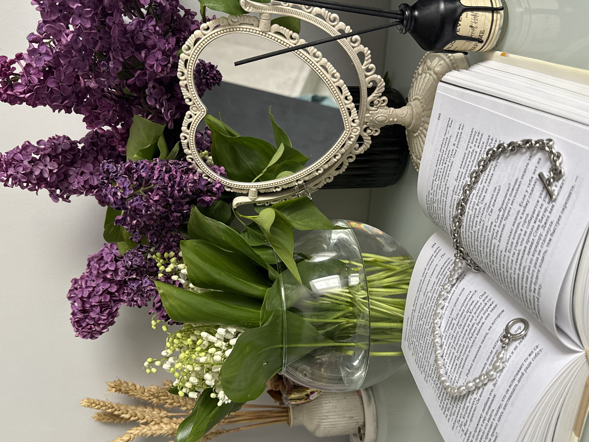 A still life composition captures a vibrant bouquet of purple lilac and white lily of the valley flowers with prominent green leaves, arranged in a clear glass vase. An ornate, heart-shaped ivory mirror with intricate detailing stands behind the flowers. To the right, an open book with Russian text is displayed, featuring page numbers 284 and 285. Adorning the book are a silver chain bracelet with a cross pendant and a pearl bracelet with a silver ring clasp. In the background, a dark reed diffuser bottle with black reeds is partially visible, along with an off-white decorative object or stand. Dried wheat stalks are visible at the bottom left. The scene is brightly lit, suggesting an indoor daytime setting, with a plain light-colored background. Reflections in the mirror indicate a window or light source. Visible text on the book includes Russian words and names like "Кэтрин", "Рейчел", "Джек", and "Сан-Франциско", while labels on the diffuser bottle show English words such as "ENJOY", "EVE", "Key", "riott Hotel", and "PERFUME".
