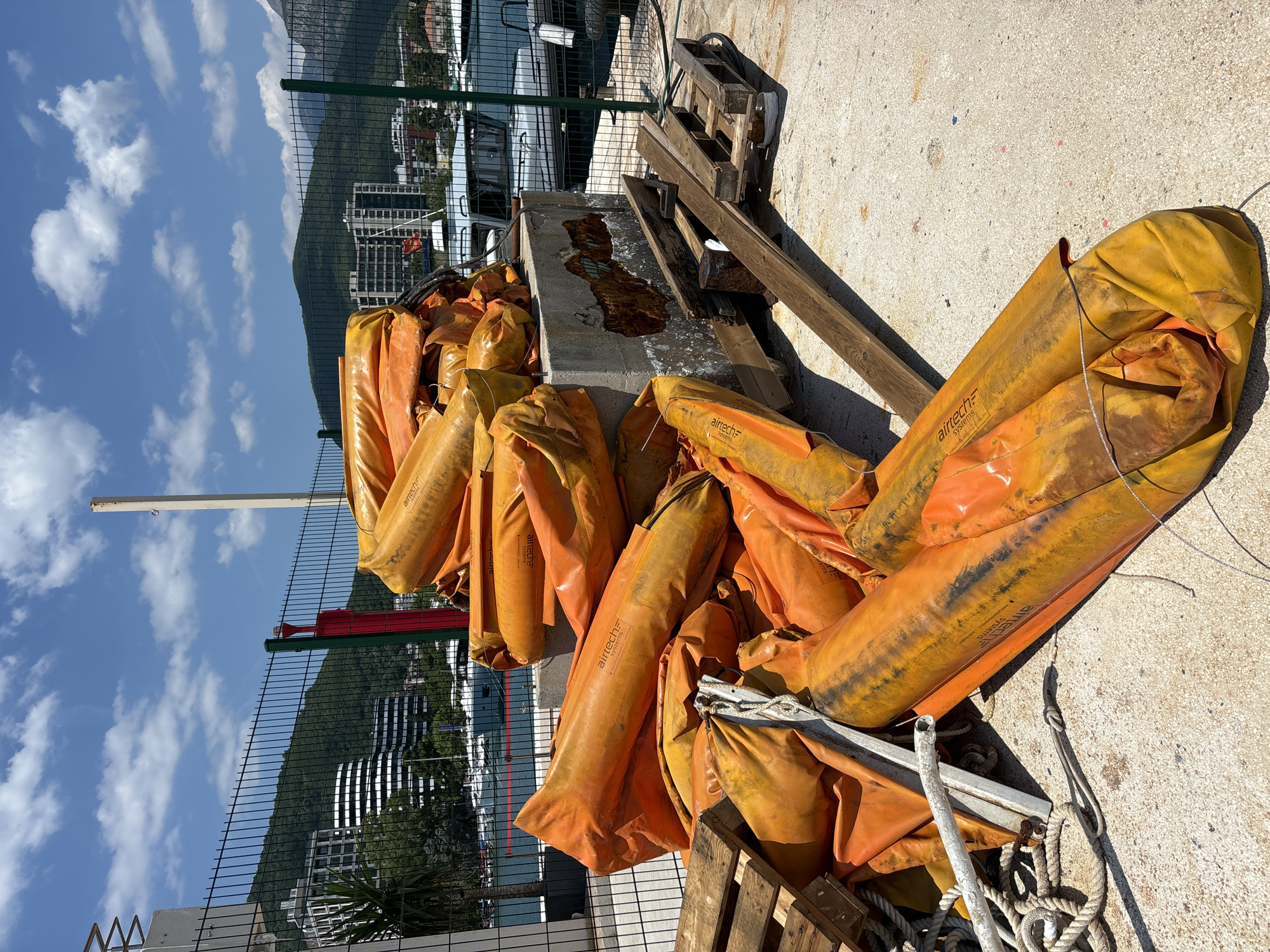 The image shows a pile of deflated orange and yellow inflatable objects, possibly buoyancy aids or barriers, stacked on a concrete surface next to a body of water. In the background, a green metal fence separates the foreground from a scene of buildings and lush green hills under a bright blue sky with scattered white clouds. The time of day appears to be daytime, likely sunny, given the clear sky and shadows. Some of the inflatable objects have "airtech systems" visible in text. There are also wooden pallets and ropes near the pile of inflatables. The setting suggests a coastal or harbor area.