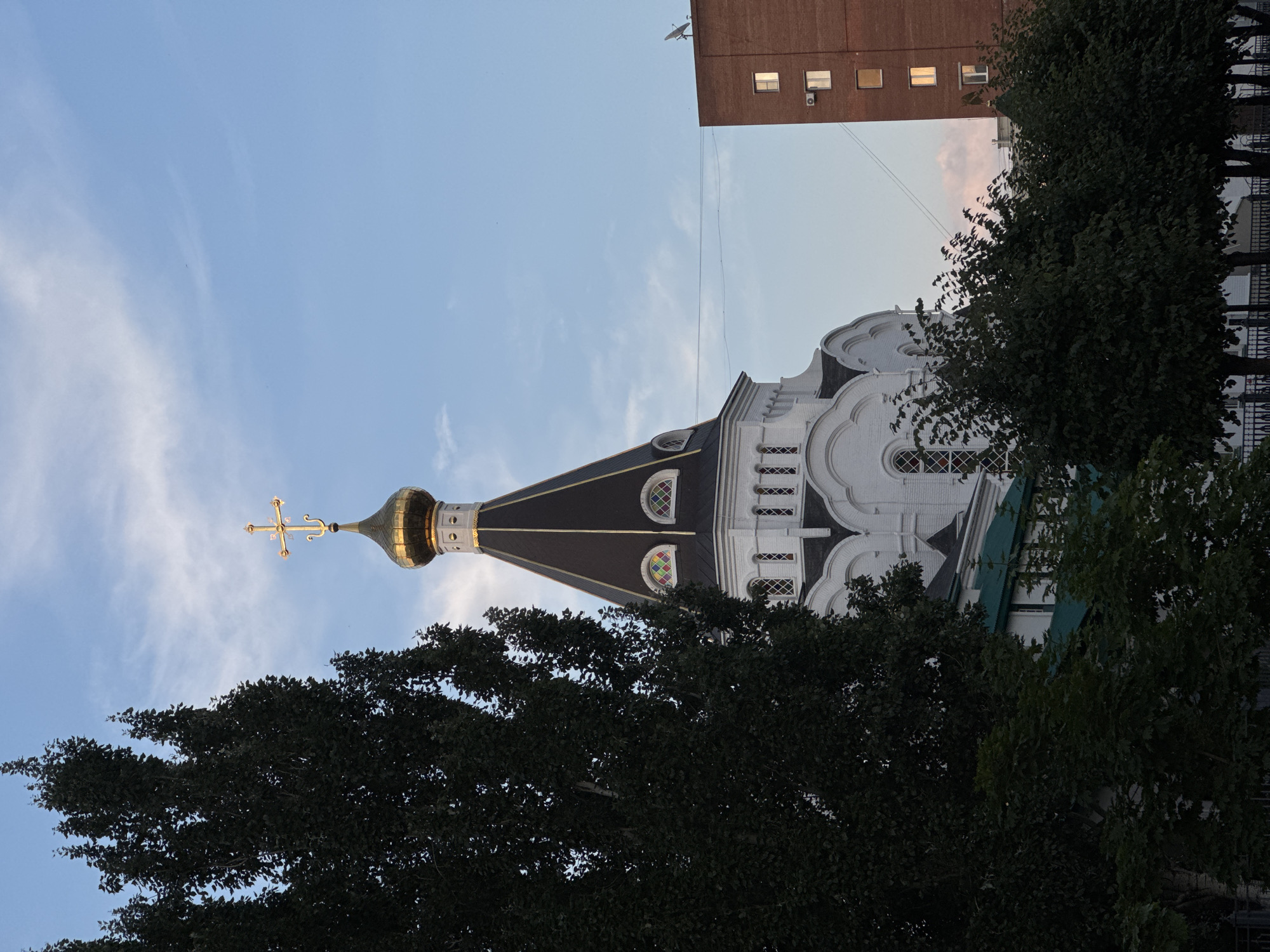 A white church with a gold dome and cross stands tall against a partly cloudy blue sky in Unknown, Unknown. The church features a dark sloped roof with decorative stained-glass windows. Adjacent to the church, a tall, dark green tree with dense foliage partially obstructs the view. To the right of the church, a red brick building with visible windows and a satellite dish is present. The lower portion of the image reveals more trees and a black metal fence.