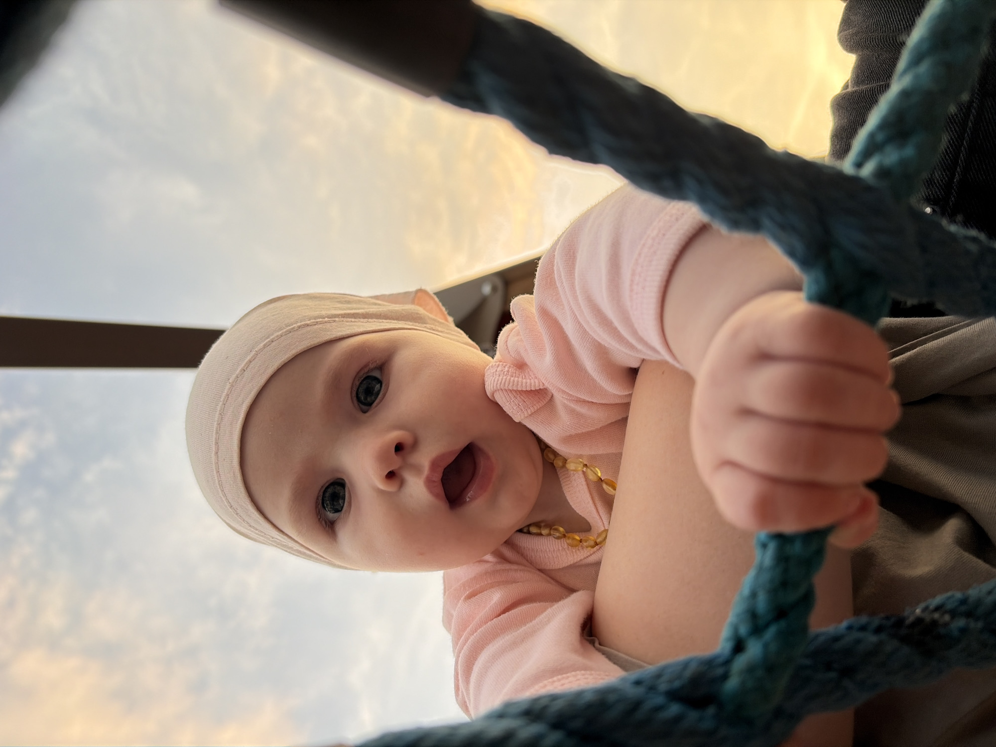 An extreme close-up, low-angle shot shows a baby looking directly at the camera with an open mouth and wide eyes. The baby is wearing a pink long-sleeved shirt, a beige head covering, and an amber bead necklace. The baby is being held by an unseen person, whose arm is visible supporting the baby. In the foreground, a thick teal rope is prominently featured, with the baby's hand grasping it. The background consists of a hazy sky with soft clouds, illuminated by the warm tones of sunset. The setting appears to be an outdoor recreational area, possibly a playground, given the presence of the rope. The image does not contain any recognizable elements that would allow for precise identification of the city of Noviy, Russia.
