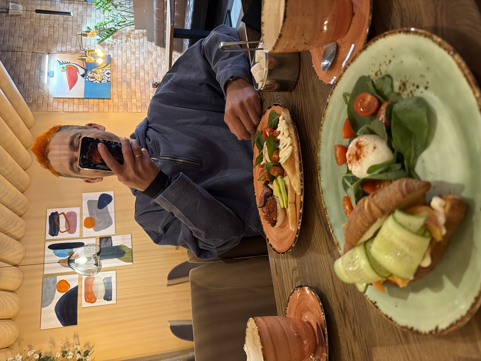 A man with orange hair is taking a photo of two plates of food on a wooden table inside a restaurant. The plate on the left has hummus, avocado, tomatoes, meat, and pita bread. The plate on the right has a croissant sandwich with salmon and cucumber on a bed of spinach and sliced cherry tomatoes, topped with a white ball of cheese. The man is wearing a blue denim jacket and holding a black smartphone. There is a container of sugar cubes nearby. In the background, there is a brick wall with paintings, a light fixture, and a plant. The location is Chaikovsky, Russia.