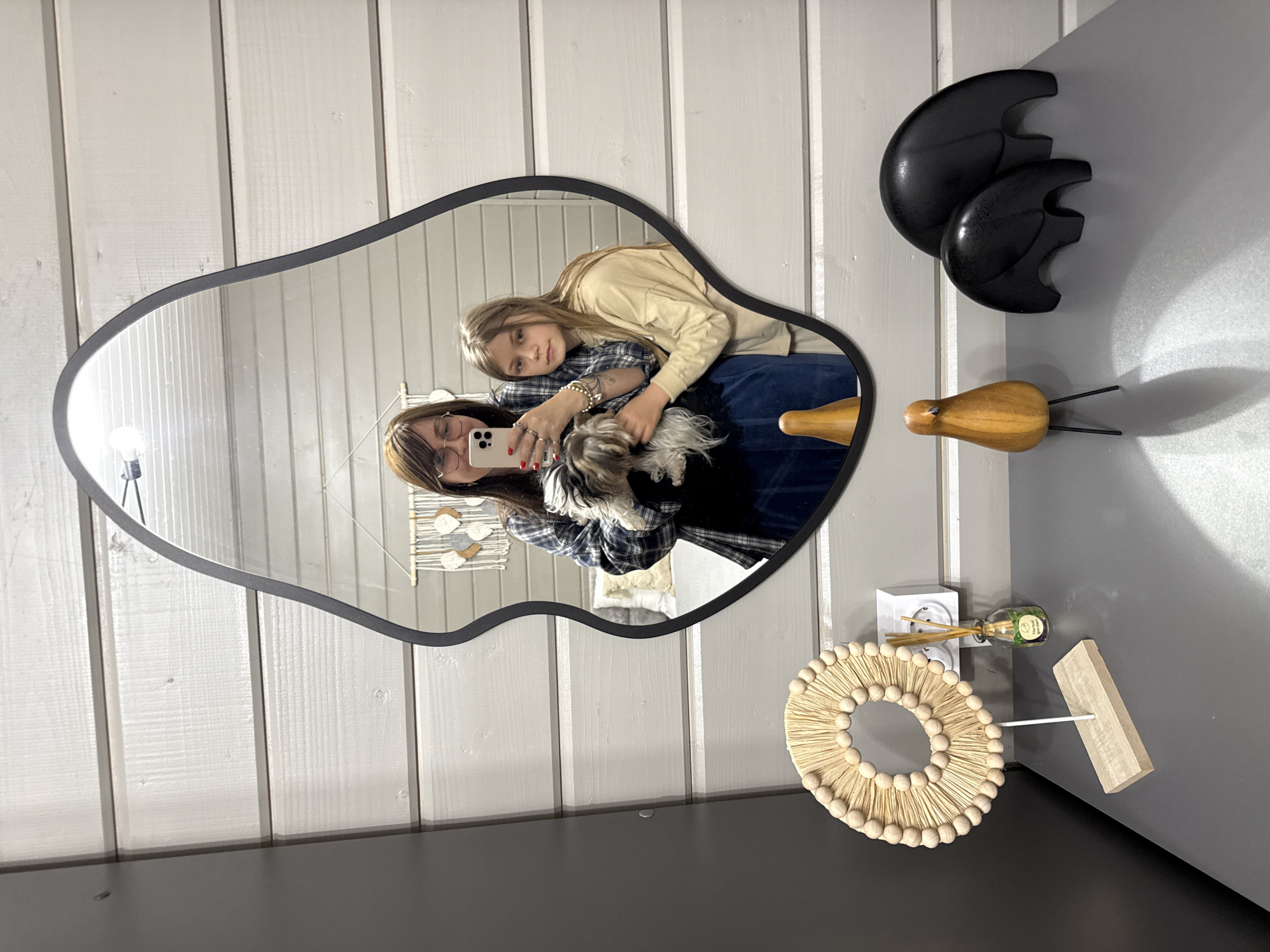 A reflection in an irregular-shaped mirror shows a woman holding a white smartphone, embracing a girl who is hugging a small dog. The woman has brown hair and wears glasses and a plaid shirt. The girl has blonde hair and wears a beige sweatshirt. The dog is a small breed with white and black fur. In the background, the mirror reflects a partially visible wall hanging with a design of birds. The wall behind the mirror is white with vertical wood paneling. Adjacent to the mirror are black sculptures resembling abstract crescent shapes, a wooden bird figurine with wire legs, a white outlet with diffuser sticks, a diffuser bottle, and a wooden stand with a straw wreath. The location is suggested by the fact that the person who made the request is located in Buzhaninovo, Russia.
