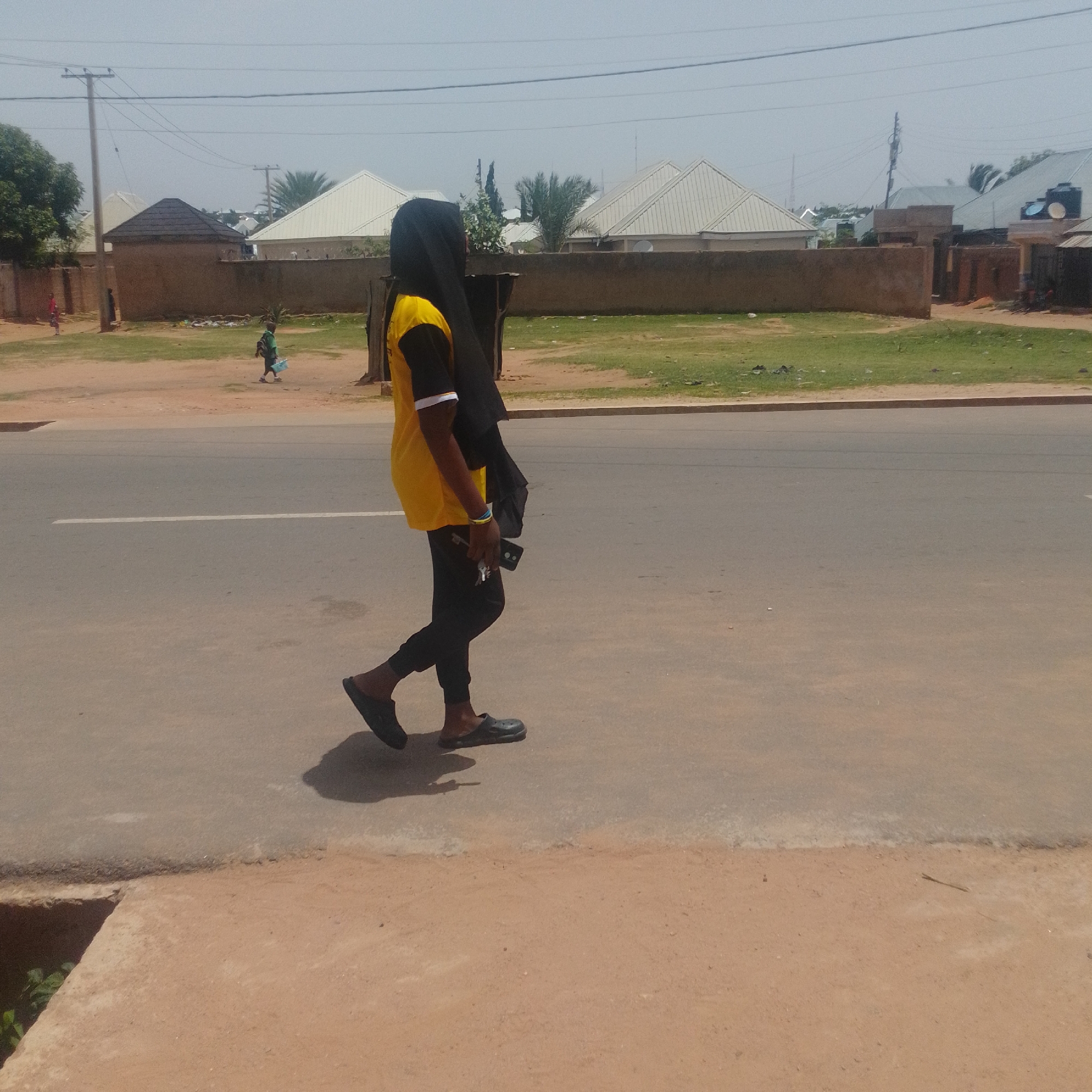 A young woman wearing a black hijab and a yellow and black shirt is walking on the side of a paved road in Gombe, Nigeria. She is wearing black pants and black slip-on shoes. She is carrying a black mobile phone and a set of keys in her right hand. To the left of the road is a strip of green grass, and behind that is a low, brown wall with several buildings and white-roofed structures visible. A utility pole with power lines stands near the wall. In the background, there are palm trees and other vegetation. The sky is clear and overcast. Another person, a child, is visible in the distance walking on the grassy area.