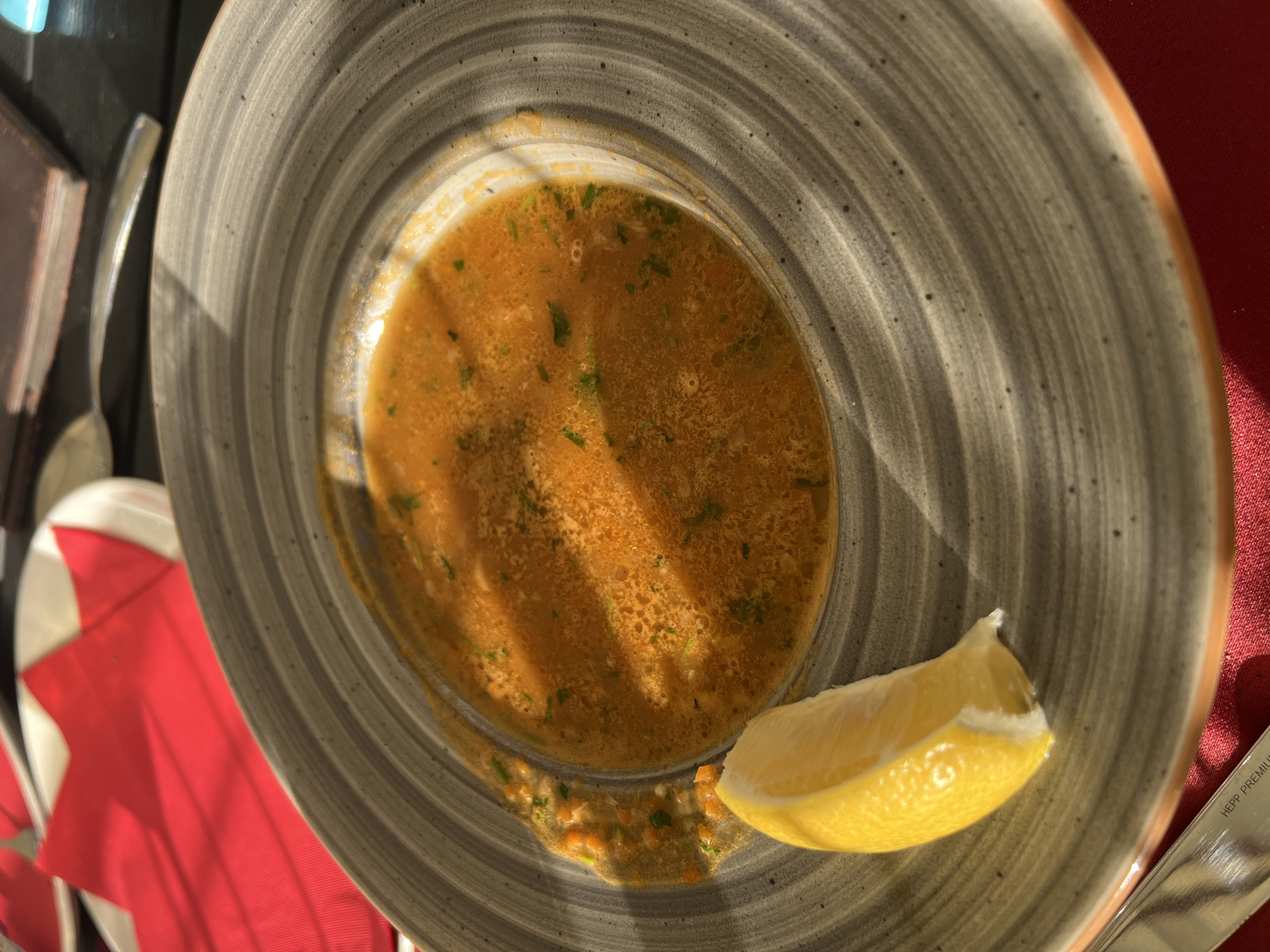 A close-up, overhead shot features a bowl of orange soup with visible herbs and a lemon wedge on the side, placed on a grey, textured plate. The plate is on a dark surface, likely a table, with a red cloth underneath. In the background, a red napkin is folded on a white plate. A spoon rests on the table next to a stack of what appears to be menus. The scene is lit by natural light, casting shadows. The setting is a restaurant in Sveti Stefan, Montenegro.