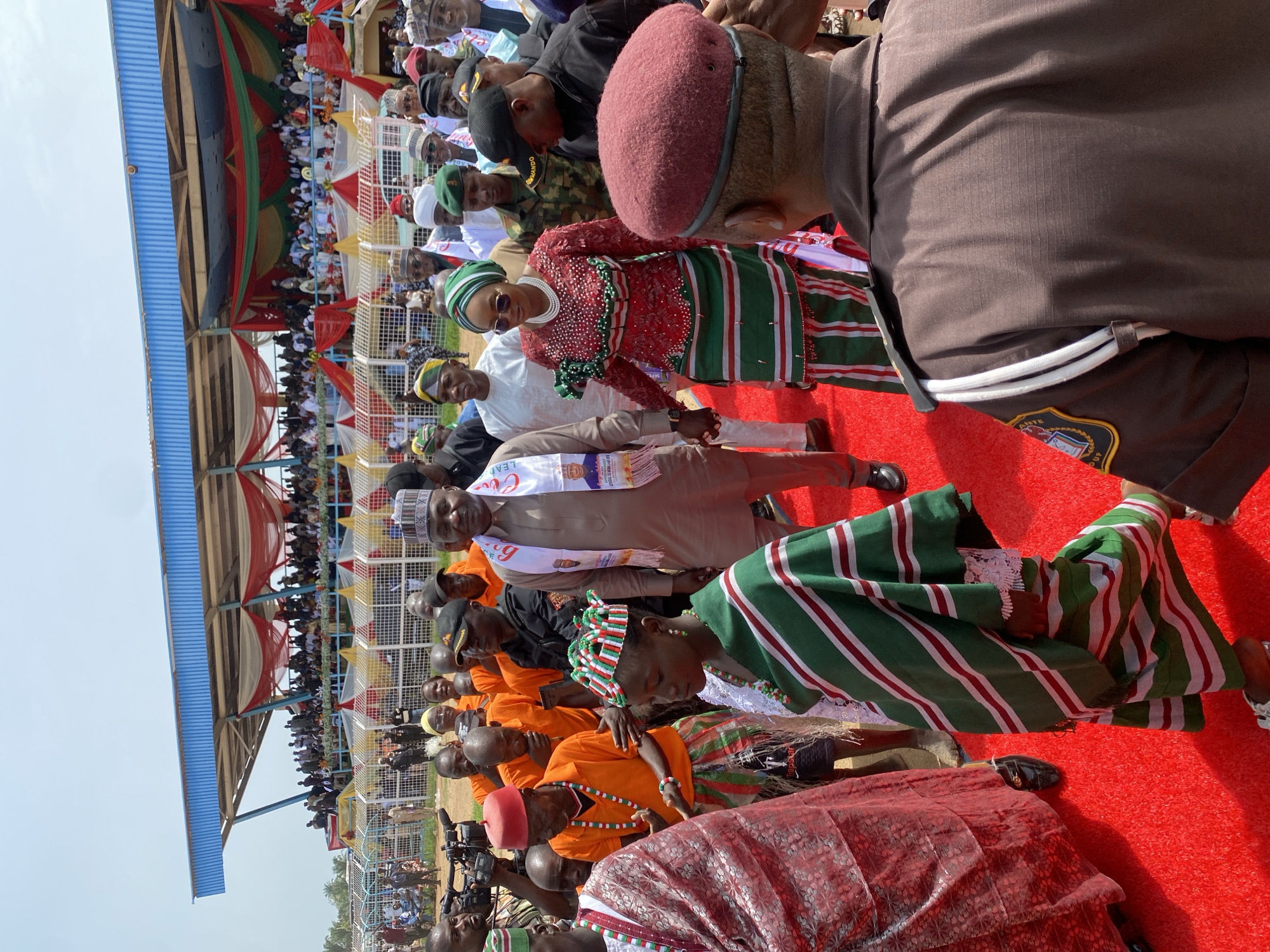 The image shows a crowded outdoor event in what is possibly Maigizo, Nigeria. A large crowd of people is visible in the stands of a stadium or event venue. In the foreground, several individuals stand on a red carpet. Many of the people are wearing traditional clothing. Several people are dressed in orange shirts with orange necklaces, while others are wearing red and green striped fabrics. Some of the crowd is covered by a tent-like structure. The sky is clear and bright.