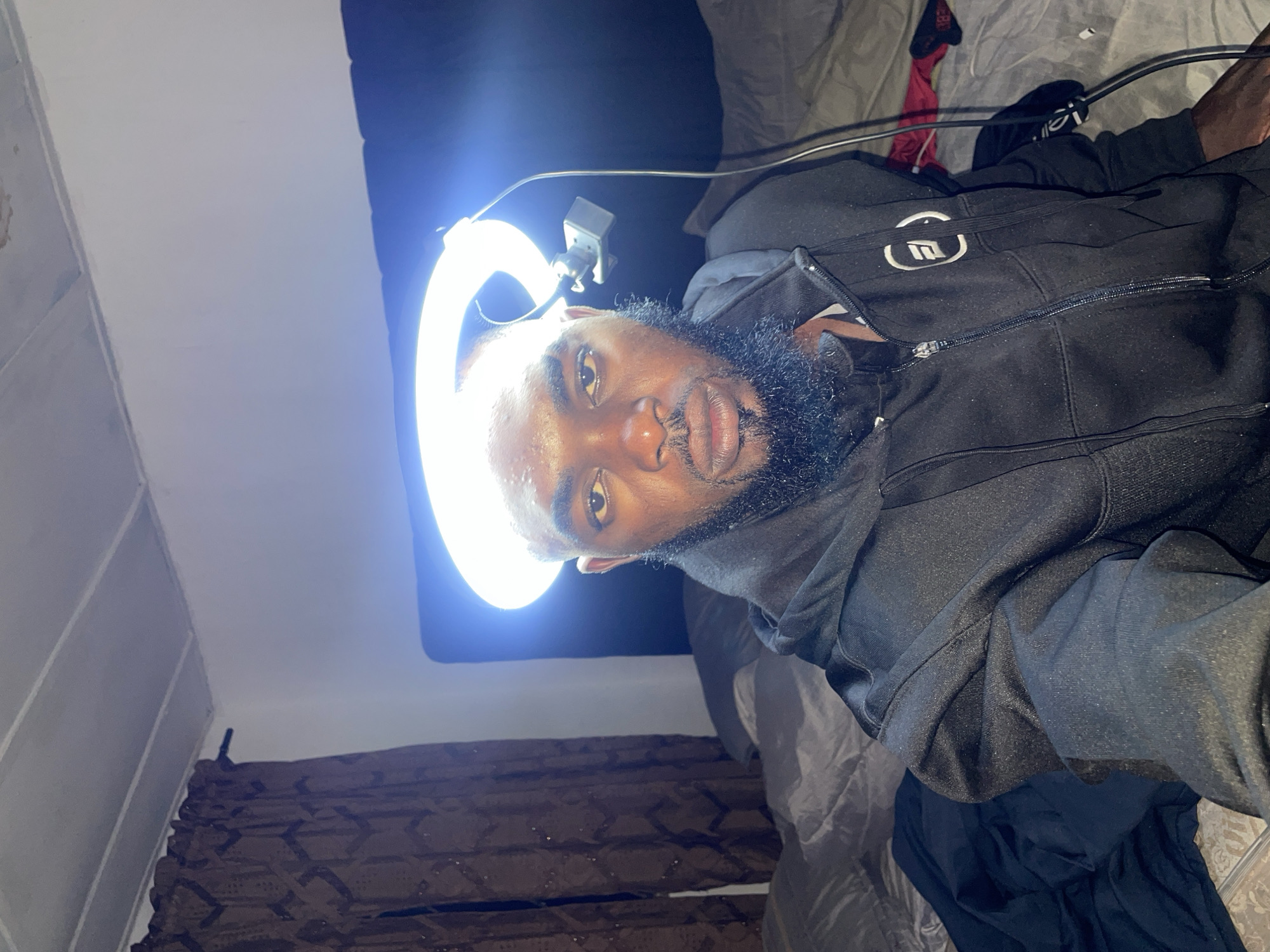 This is a matter-of-fact description of an indoor scene in Jos, Nigeria.
An adult male, positioned centrally, is taking a selfie. He has a dark complexion, a full beard, and short hair on his head, with visible perspiration on his forehead and nose. He is wearing a black zippered hoodie with a white, circular, abstract logo on the left chest/shoulder area. Directly above his head, a bright LED ring light serves as the primary illumination source, creating a prominent halo effect and casting strong light onto his face. Wires from the light extend downwards on the right side of the frame.
The background shows a bedroom environment. A large bed with rumpled dark grey bedding (sheets, pillows, comforter) occupies the mid-ground. A dark headboard is visible behind the man. To the left, dark brown patterned curtains are drawn, covering a window, with a dark curtain rod visible at the top. The walls are light-colored, possibly white, and the white ceiling above features parallel structural lines or panels. Various items of dark and red clothing are scattered on the bed and floor to the right side of the frame.