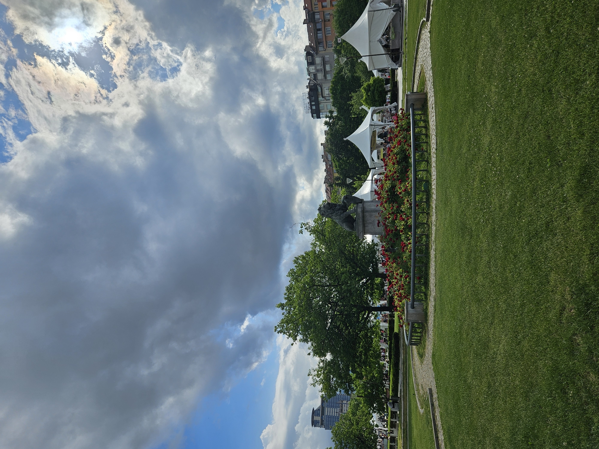 This image captures a vibrant outdoor scene within a public park or garden in Sofia, Bulgaria, under a dynamic, cloudy sky. The sun, partially obscured by clouds, casts bright light from the upper left, creating a dramatic effect.
To the right, a wide expanse of neatly maintained green grass stretches across the foreground and midground, bordered by a curving paved path made of cobblestones or similar material. Along the left side of this path, several large, white canopy tents are set up, indicating an outdoor cafe or restaurant. Under these tents, numerous individuals are visible, seated at tables and appearing to enjoy a meal or drinks. The word "MANGIA" is clearly visible in dark lettering on the side of one of the white tents.
Further to the left, a dense line of lush green trees provides shade and depth. A decorative arrangement of vibrant red roses or flowering bushes lines a fence or barrier, adding a splash of color. Amidst the greenery and flowers, a stone statue, possibly a lion, stands on a pedestal. In the background, urban buildings with varied architecture are visible, hinting at the city surroundings. The sky is a mix of bright white and darker grey clouds, with patches of blue sky discernible, particularly in the lower left. The overall impression is one of a bustling yet relaxed urban park, with people enjoying the outdoor amenities on a partly cloudy day.