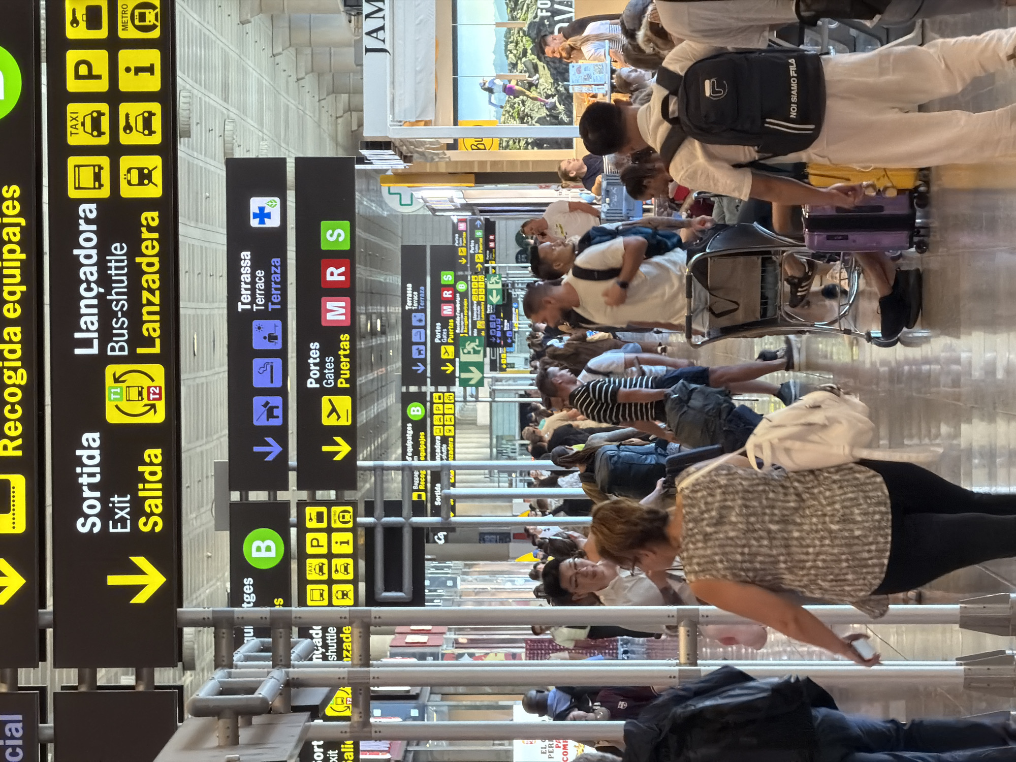 The image depicts a bustling interior scene at El Prat Airport in El Prat de Llobregat, Spain, likely within the arrivals or departures hall. Numerous passengers, identifiable by their varied attire, luggage, and backpacks, are navigating the space. The environment is characterized by modern airport architecture, including polished floors, metal pillars, and prominent signage. The signage, in multiple languages (Spanish, Catalan, English, and potentially French), indicates directions for various airport services, such as "Sortida" (Exit), "Salida" (Exit), "Llançadora" (Bus-shuttle), "Lanzadera" (Bus-shuttle), "Terrassa" (Terrace), "Portes" (Gates), and "Puertas" (Gates). Icons on the signs represent transportation options like taxis, buses, cars, metro, and baggage claim. Some individuals are seen with rolling suitcases and airport trolleys. The lighting appears to be a mix of ambient indoor lighting and overhead illumination, contributing to a well-lit but somewhat diffused atmosphere. The overall impression is one of a busy transit hub with travelers in motion.