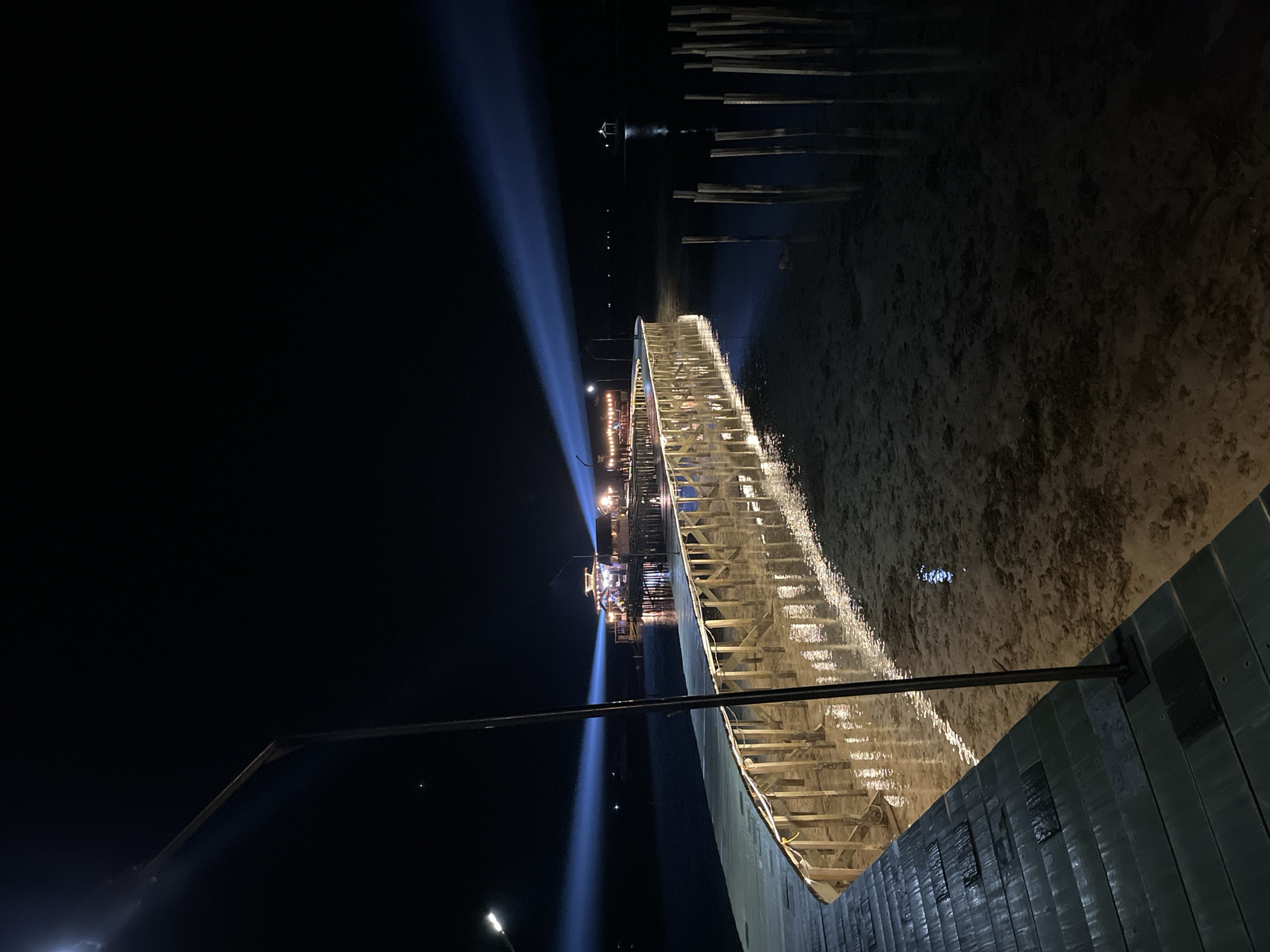 The image captures a nighttime view of a wooden pier extending over shallow water near Karimunjawa, Indonesia. The pier is illuminated by string lights and a street lamp, casting reflections on the water's surface. In the distance, a building or structure on the pier is brightly lit, and powerful blue spotlights are projected into the dark sky from this structure. The water appears calm, with the sandy bottom visible in places. Several vertical wooden posts are submerged in the water to the right of the pier. The overall scene suggests a lively atmosphere, possibly a tourist destination or event venue.