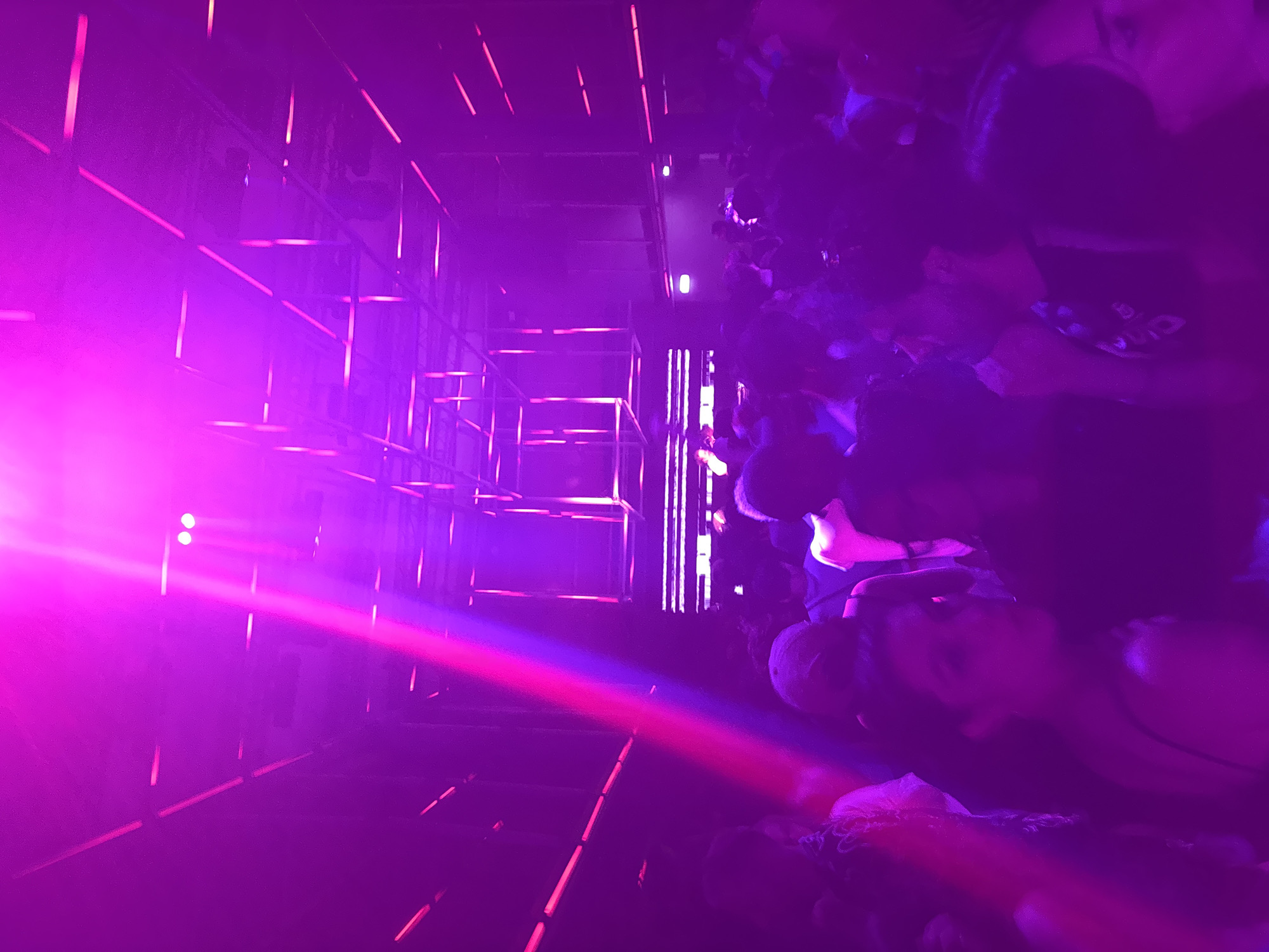 Captured at an angle, the image depicts a dimly lit scene, likely a nightclub or event space, saturated with a vibrant purple hue. A geometric grid-like structure stands on the left side, illuminated with the same purple light. In the distance, a bright light source is visible. A crowd of people occupies the right side of the frame, their features obscured by the darkness. The location is not specified, and there is no clear indication that it is in Paris, France.