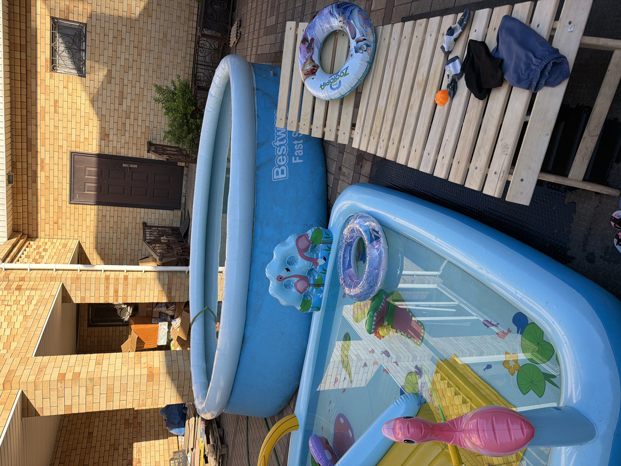 The image shows a backyard scene with two inflatable swimming pools. In the foreground, a large blue inflatable pool is filled with water and contains various inflatable toys, including a yellow slide, a pink flamingo, and a purple ring. A smaller inflatable pool is visible behind it, also filled with water and containing a light blue inflatable ring with flamingos printed on it. To the right of the pools, a wooden plank structure is laid out, with several items placed on it: a pair of socks, a black piece of clothing, a blue garment, and an orange ball. The scene appears to be set outdoors on a sunny day, as indicated by the lighting and shadows. The background shows a brick building with a door and a window, suggesting a residential setting. The image is taken from a slightly elevated angle, looking down on the pools and surrounding area. There is no visible text in the image. The overall impression is one of a recreational setup for a warm day.