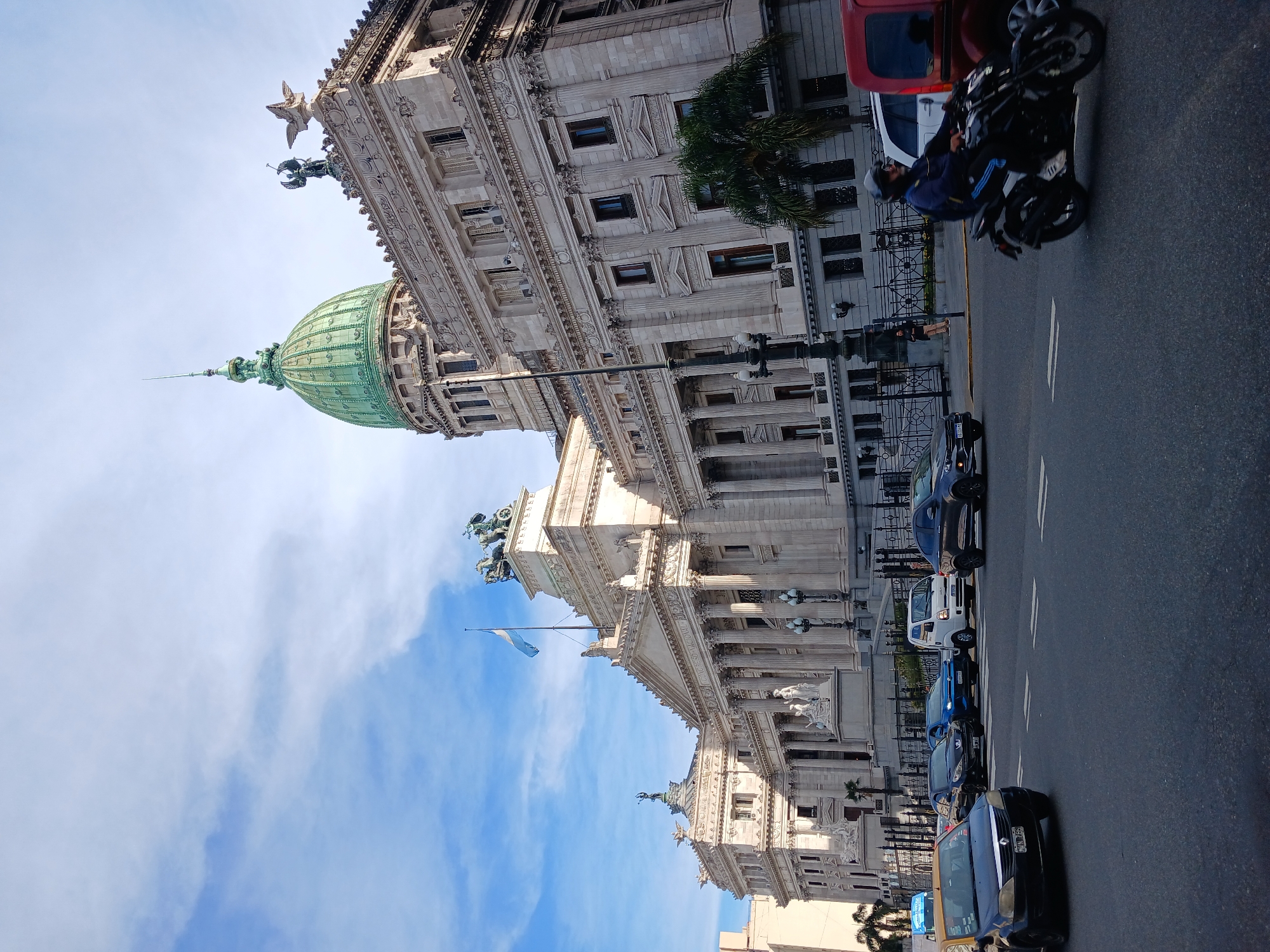 The image shows the Palace of the Argentine National Congress in Buenos Aires, Argentina. The building, constructed of light-colored stone, features a green dome, classical columns, statues, and ornate detailing. An Argentine flag is flying. Street-level details include several parked vehicles, a motorcycle with a rider, a person standing on the sidewalk, and a palm tree. The sky is blue with some clouds.