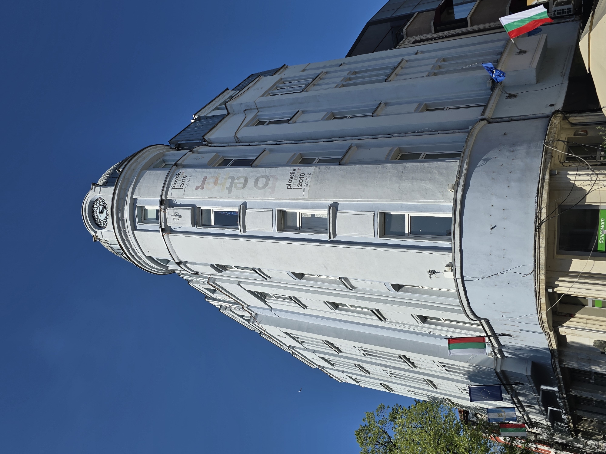 The image depicts a tall, white building in an urban environment. The building features a rounded corner section with a clock near the top and several rows of windows along its height. Banners or signs with the text "Plovdiv 2019" are visible on the curved facade. Flags, including what appears to be the Bulgarian flag, are mounted on the building. The sky is clear and blue. The building is situated in an unknown city.