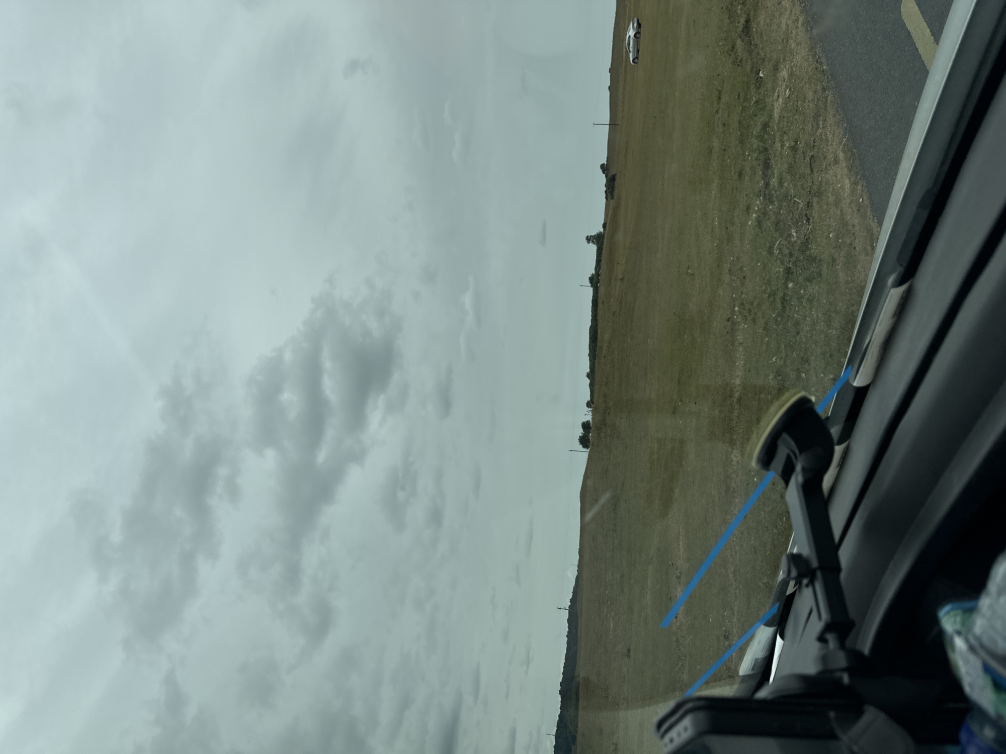 The image depicts a view from inside a vehicle, looking out at a rural landscape under a cloudy sky. The foreground is dominated by the dashboard and windshield of the car, with a dark textured surface. A black mount with a suction cup is visible on the windshield, holding a device. Two thin blue lines, possibly reflections or markings on the windshield, extend diagonally across the scene.
The middle ground features a vast, undulating grassy field with patches of dry, straw-colored grass and some darker green areas. A few scattered trees and some shrubbery dot the landscape, with power poles and lines visible in the distance.
In the far background, a low hill rises, covered in greenery and trees. The sky is overcast with a thick layer of grey clouds. To the right of the frame, on the grassy field, a silver sedan is parked. The scene appears to be set in Lumville, Ireland, a rural area characterized by its open fields and rolling hills.