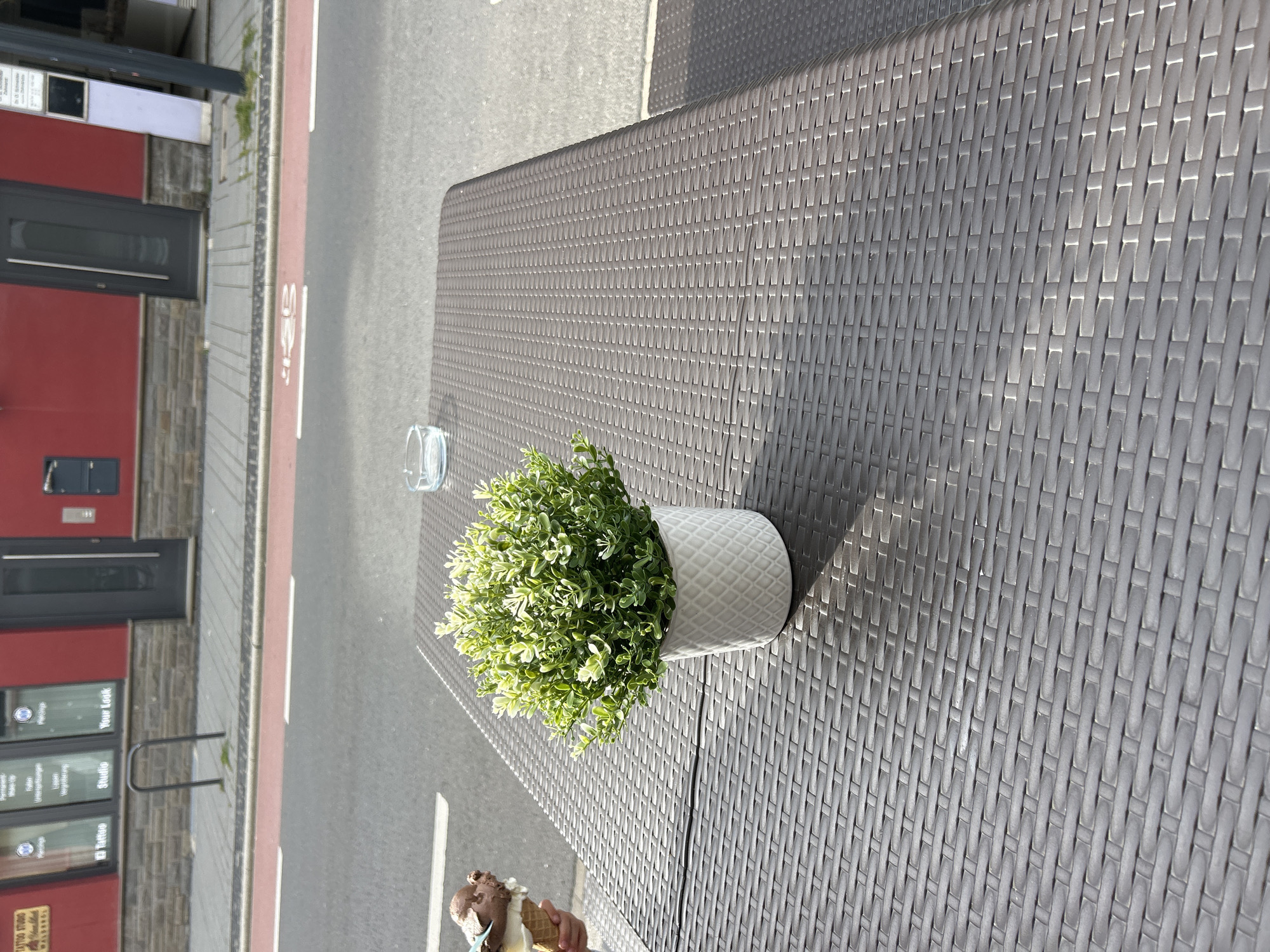 An outdoor table on a patio in Waldbröl, Germany, is partially visible. The table surface is dark brown and textured like wicker. On the table sits a white ceramic pot containing a small, green, bushy plant. To the left of the plant, a person's hand is visible holding an ice cream cone with two scoops of ice cream, one brown and one lighter colored. In the background, a street is visible with a red bike lane and a grey sidewalk. Beyond the sidewalk are buildings with red and grey facades, and a dark gray door. An ashtray with a small amount of water is also on the table.