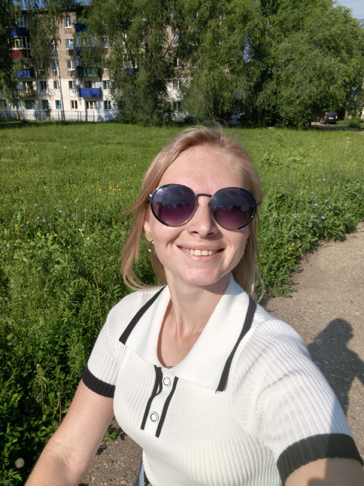 A young Caucasian woman with blonde hair and sunglasses smiles at the camera in a selfie. She is wearing a white collared shirt with black trim. The background shows a grassy area with trees and a multi-story building. This image was taken in Sterlitamak, Russia.