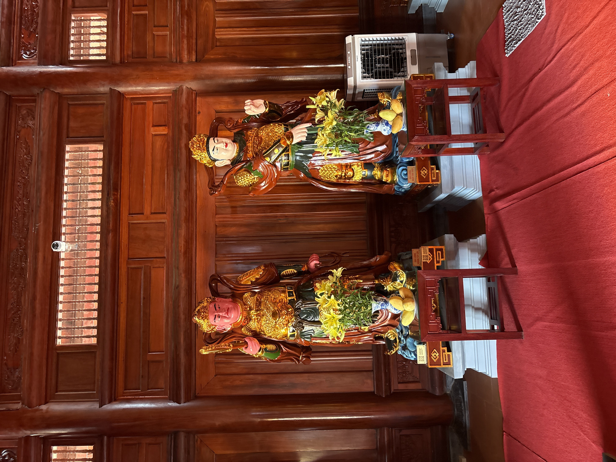The image depicts a space with ornate wooden paneling, likely within a building. Two statues of figures adorned with gold ornamentation are centrally located, each holding objects and surrounded by bouquets of yellow flowers. The statues are placed on stands made of red-lacquered wood with white bases. Next to one of the statues is a portable air cooler. The background is composed of a red fabric or rug, and a surveillance camera is visible on the wooden paneling. The image suggests a temple or other religious site in Nam Nha Trang Ward, Vietnam, given the style and features present.