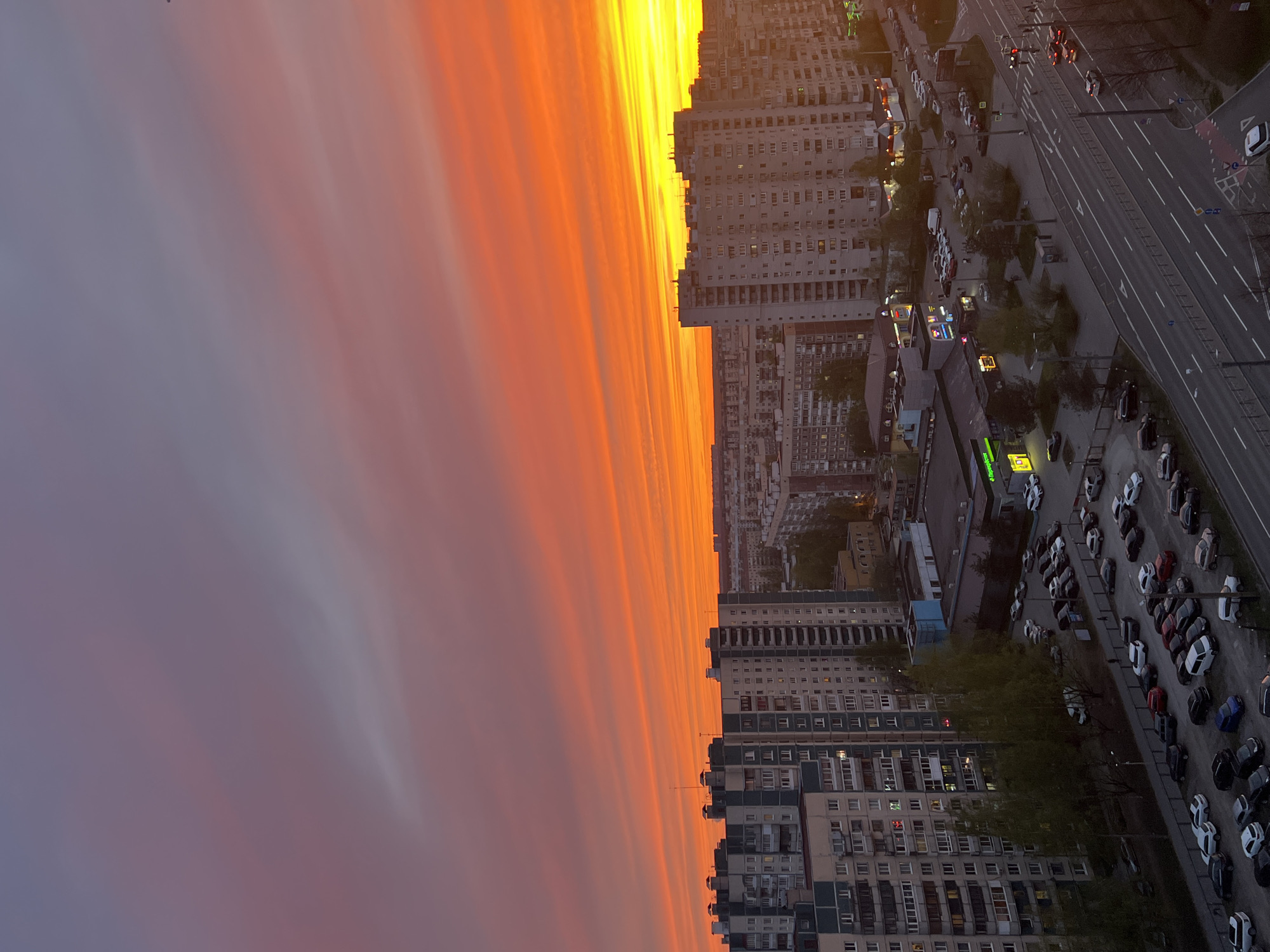 The image shows a view of a city at sunset. The sky is filled with orange and pink clouds. In the foreground, there are buildings, a parking lot filled with cars, and a street. The city is Saint Petersburg, Russia.