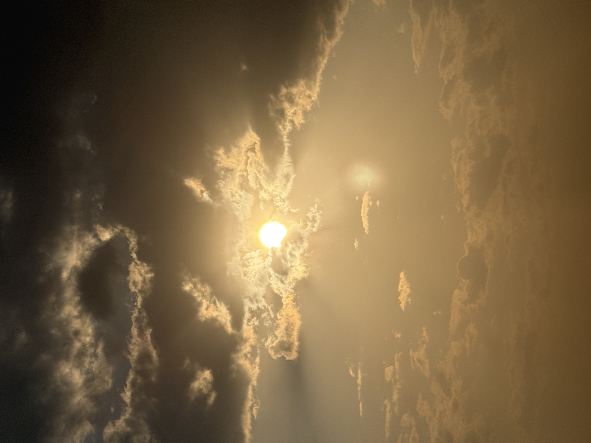 The image depicts a cloudy sky with the sun partially obscured by clouds, casting a warm, golden light. The clouds are varied in texture and density, with some appearing dark and heavy while others are illuminated by the sun's rays. The overall atmosphere suggests either sunrise or sunset. The scene is devoid of any human presence, structures, or identifiable landmarks that would pinpoint a specific location within Sary-Oy, Kyrgyzstan.